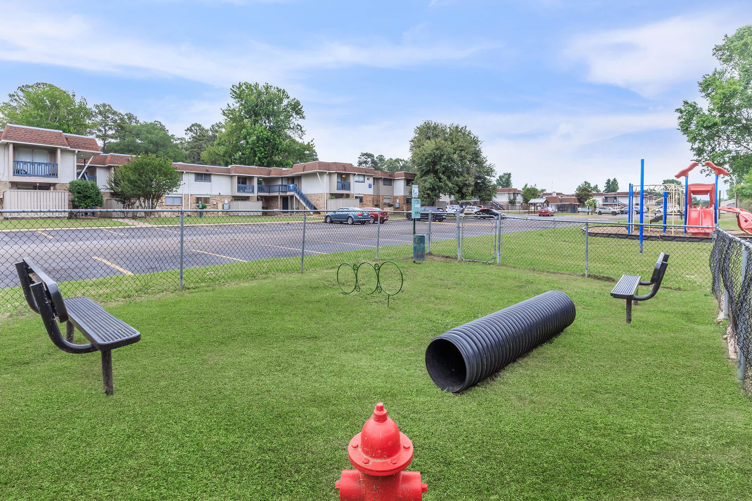 A small fenced dog park featuring green grass, two benches, a play tunnel for pets, and a red fire hydrant. In the background, there are multi-story apartment buildings and parking spaces. The scene is bright and open, ideal for pet play and relaxation.
