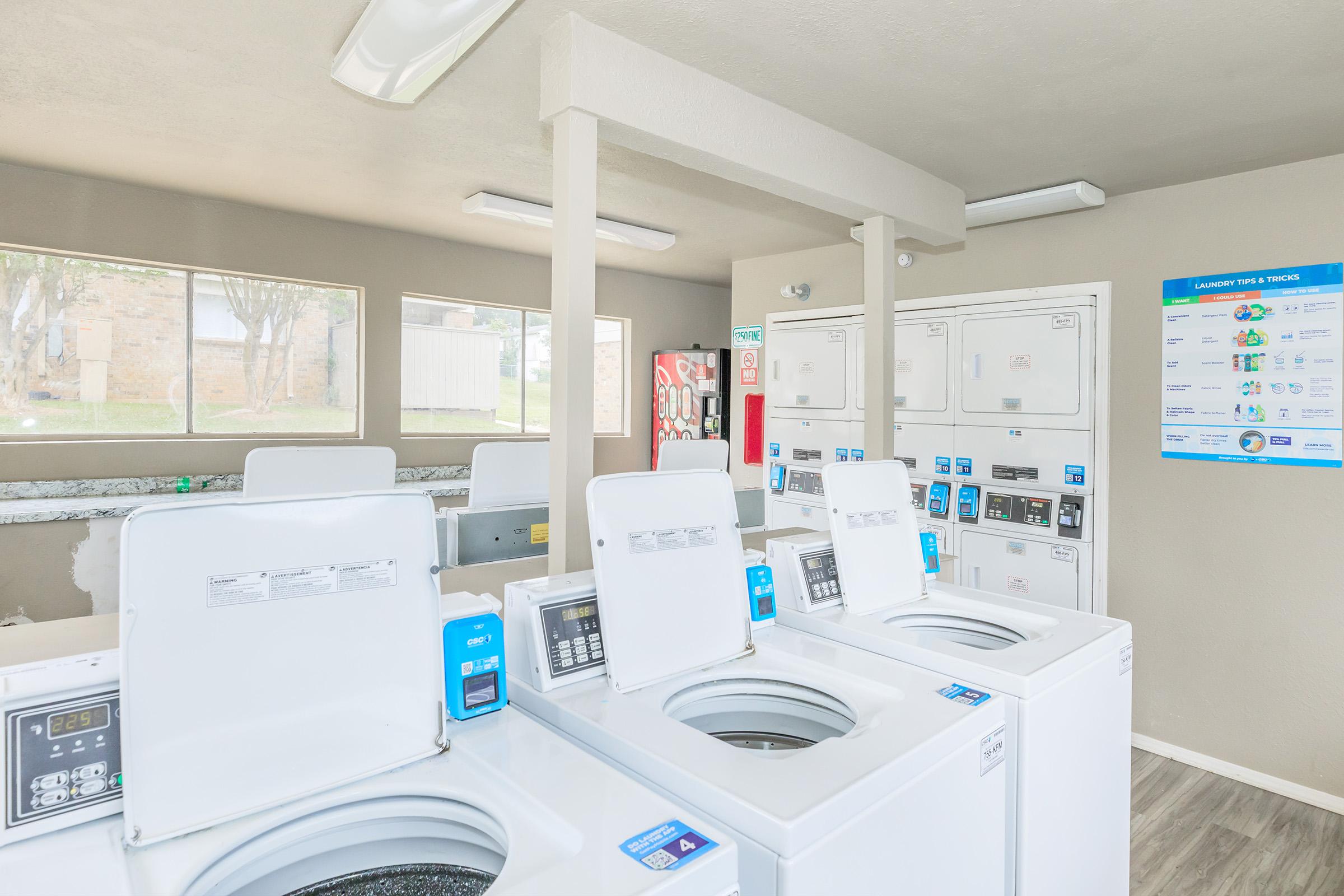 A clean, well-lit laundry room featuring several white washing machines and dryers arranged in rows. The walls are light-colored, and there’s a bulletin board with laundry guidelines on one wall. Natural light filters in through windows, creating a bright and inviting atmosphere.