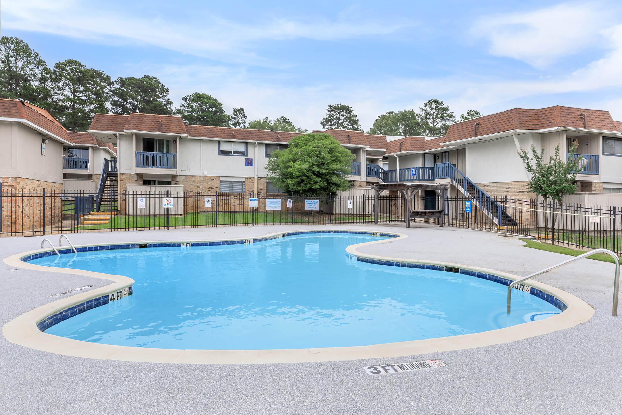 A spacious outdoor swimming pool with a wavy shape, surrounded by a fenced area. In the background, there are two-story apartment buildings with balconies and green lawn areas. The sky is clear with some clouds, creating a bright and inviting atmosphere.
