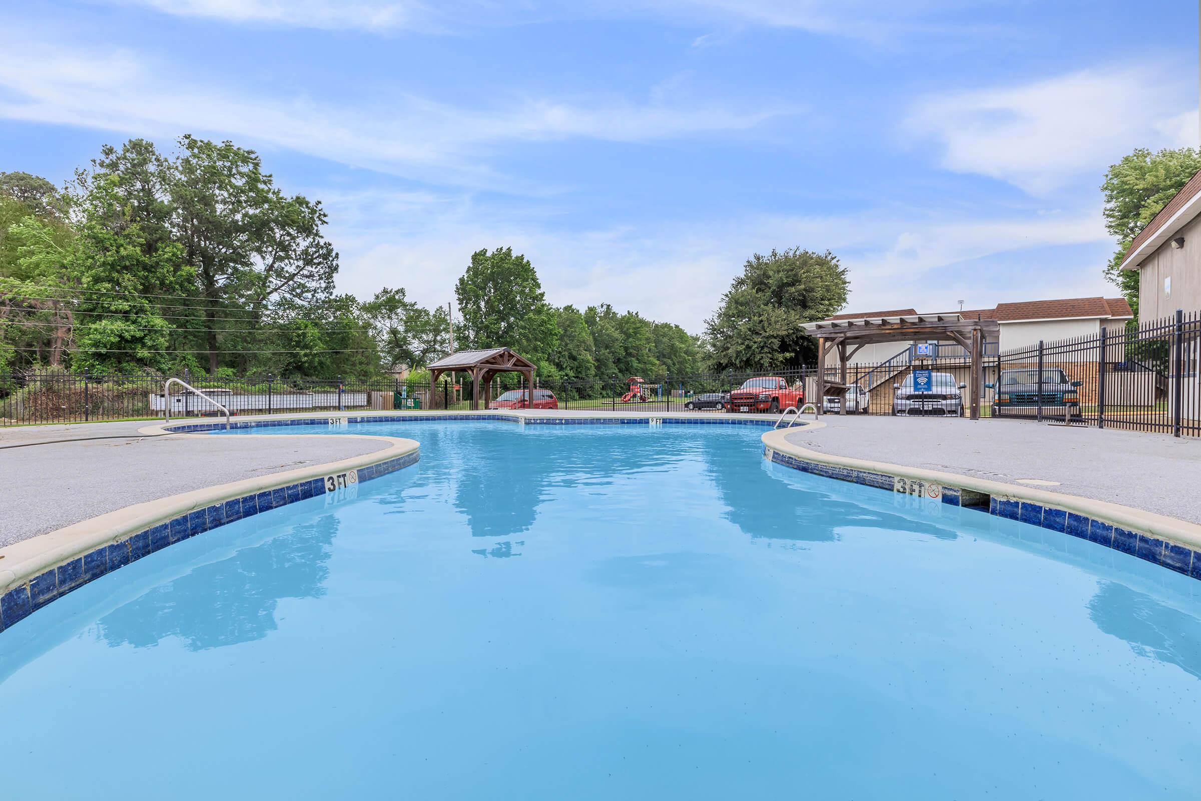 A clear blue swimming pool with a gently sloping edge, surrounded by a fence and greenery. In the background, there are trees and a building, along with a gazebo. The sky is bright with a few clouds, creating a serene outdoor atmosphere.