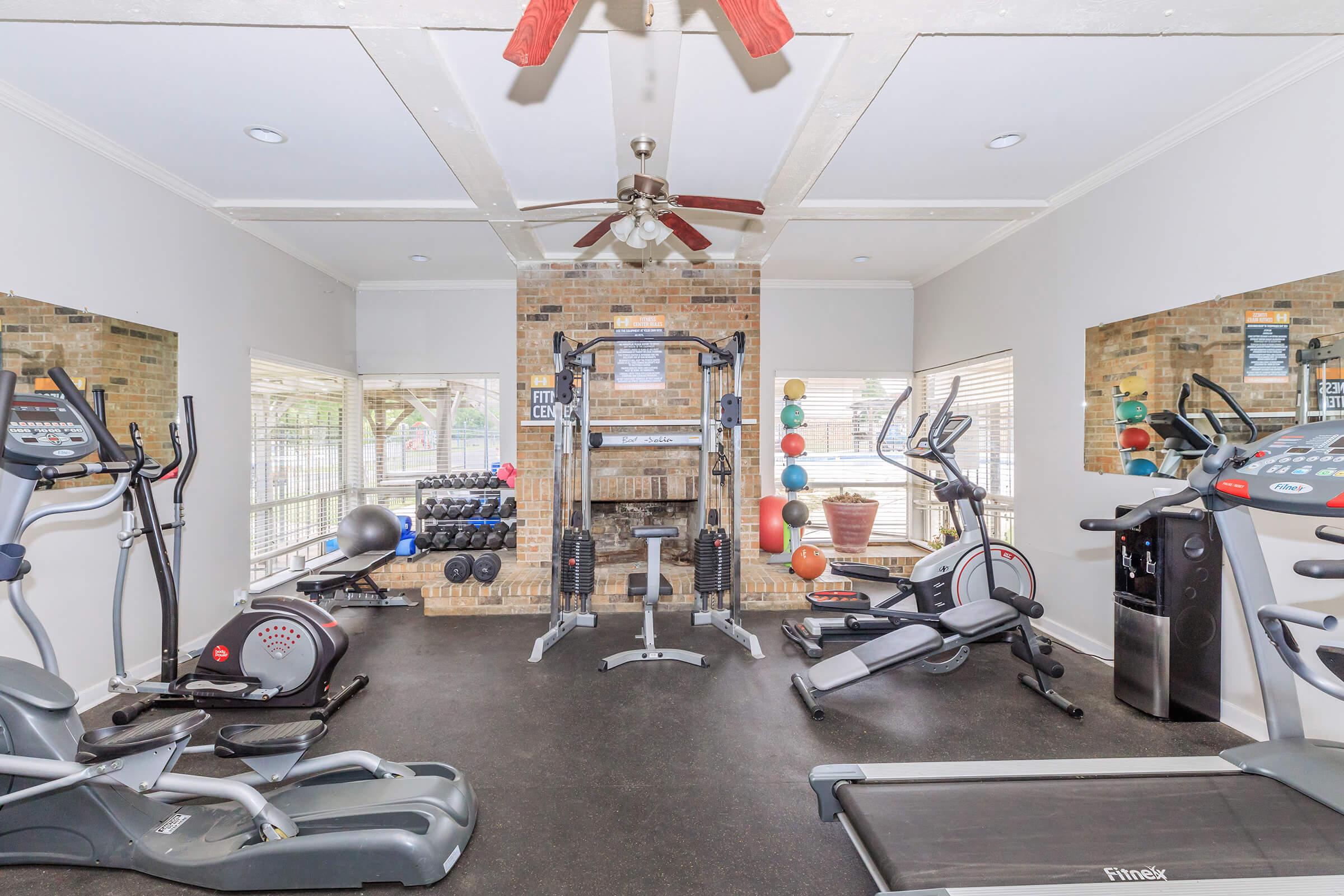 A well-equipped home gym featuring a treadmill, elliptical machine, multi-station weight machine, free weights, and exercise balls. The room is bright with large windows, brick accents, and a ceiling fan, creating a motivating workout environment.