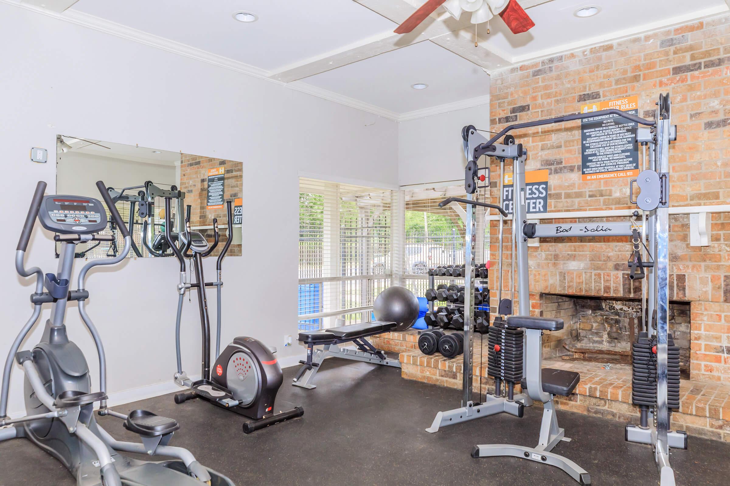 A well-equipped gym featuring a treadmill, elliptical machine, and a multi-station weight machine. It includes a bench press with weight plates, a stability ball, and free weights. The room has large windows for natural light and a brick wall design, creating a bright and inviting workout space.