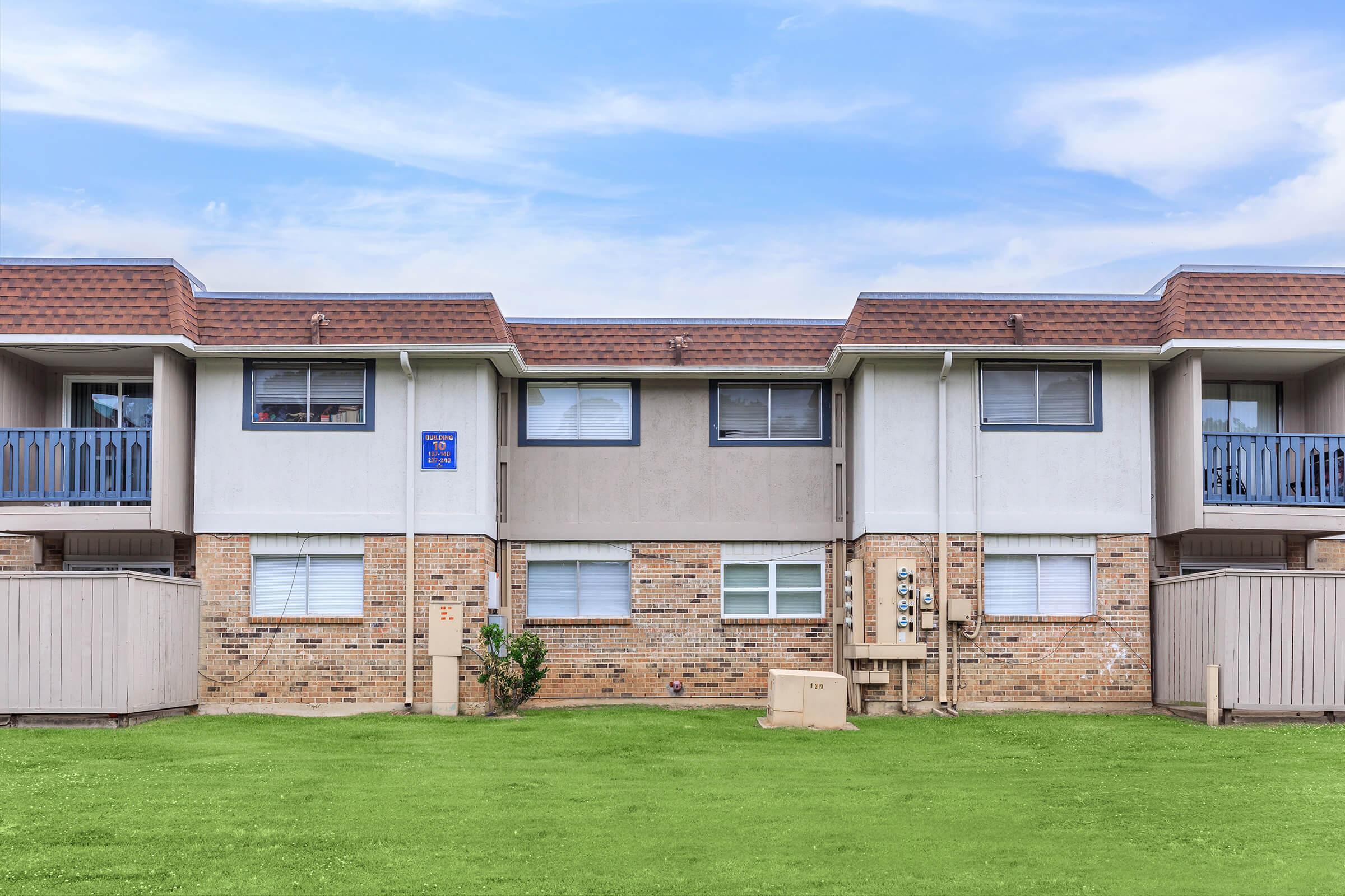 A two-story apartment building featuring a brick facade and two balconies. The ground is grassy, and there are utility meters visible on the side of the building. The sky is blue with some clouds, giving a pleasant atmosphere to the scene.