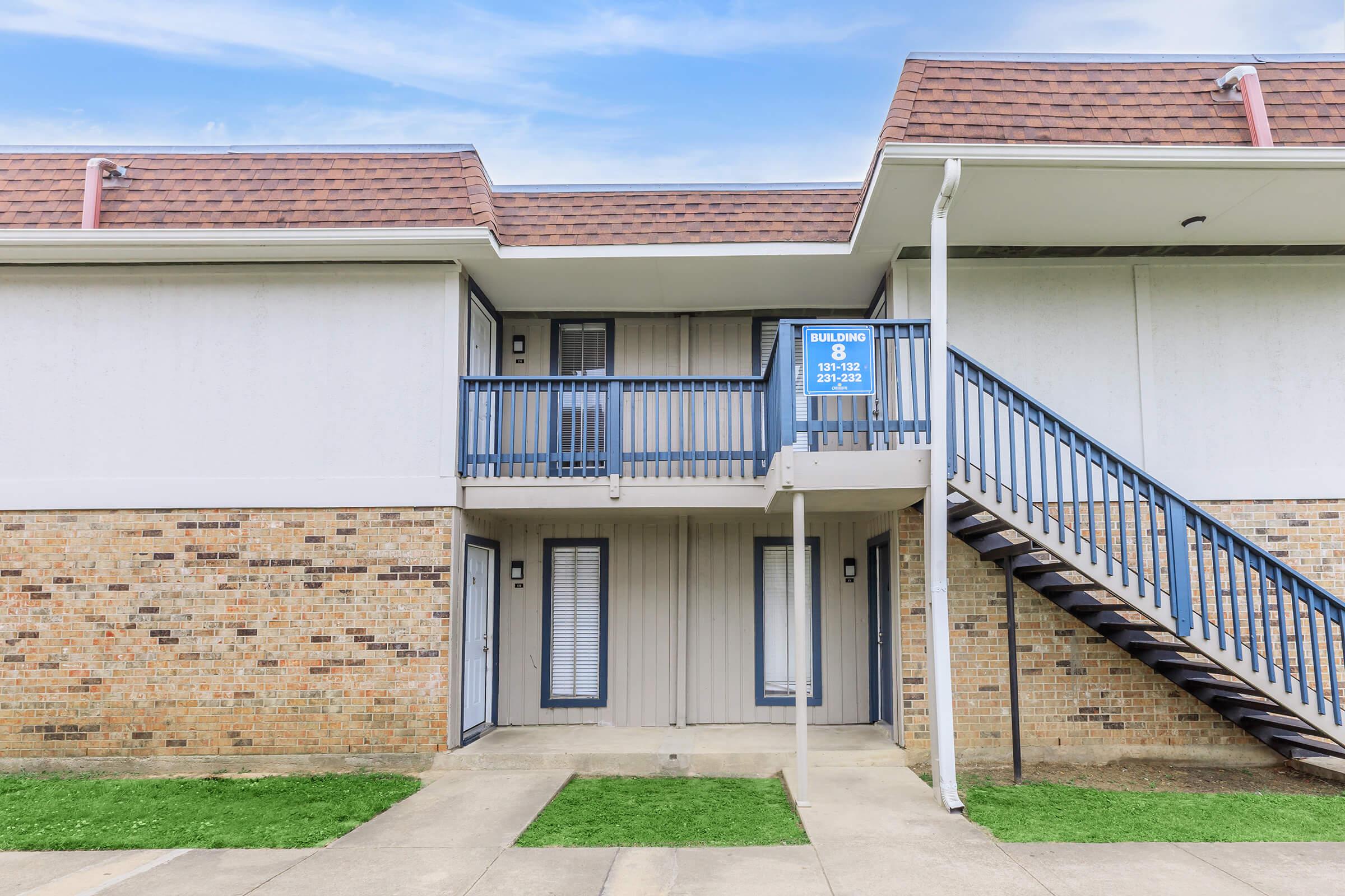 Exterior view of a two-story apartment building. The building features a brick facade and a light-colored upper section. A staircase leads to the second floor, where balconies are visible. The scene includes a clear sky and well-maintained grass in front of the entrance. A sign indicates the building number.