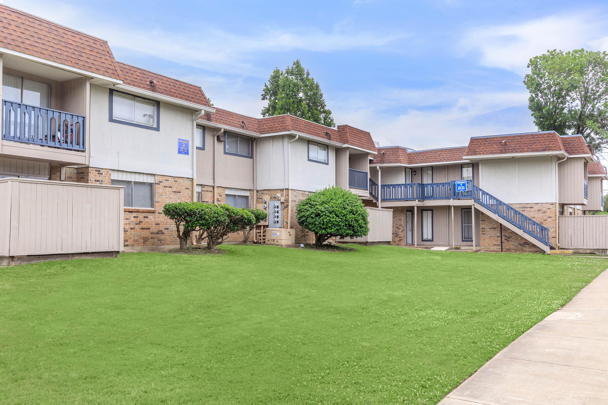 A view of a residential apartment complex featuring two-story buildings made of brick and siding. The landscape includes manicured green lawns, shrubs, and a walkway leading to the entrance of the units. The sky is blue with a few clouds, creating a pleasant atmosphere.