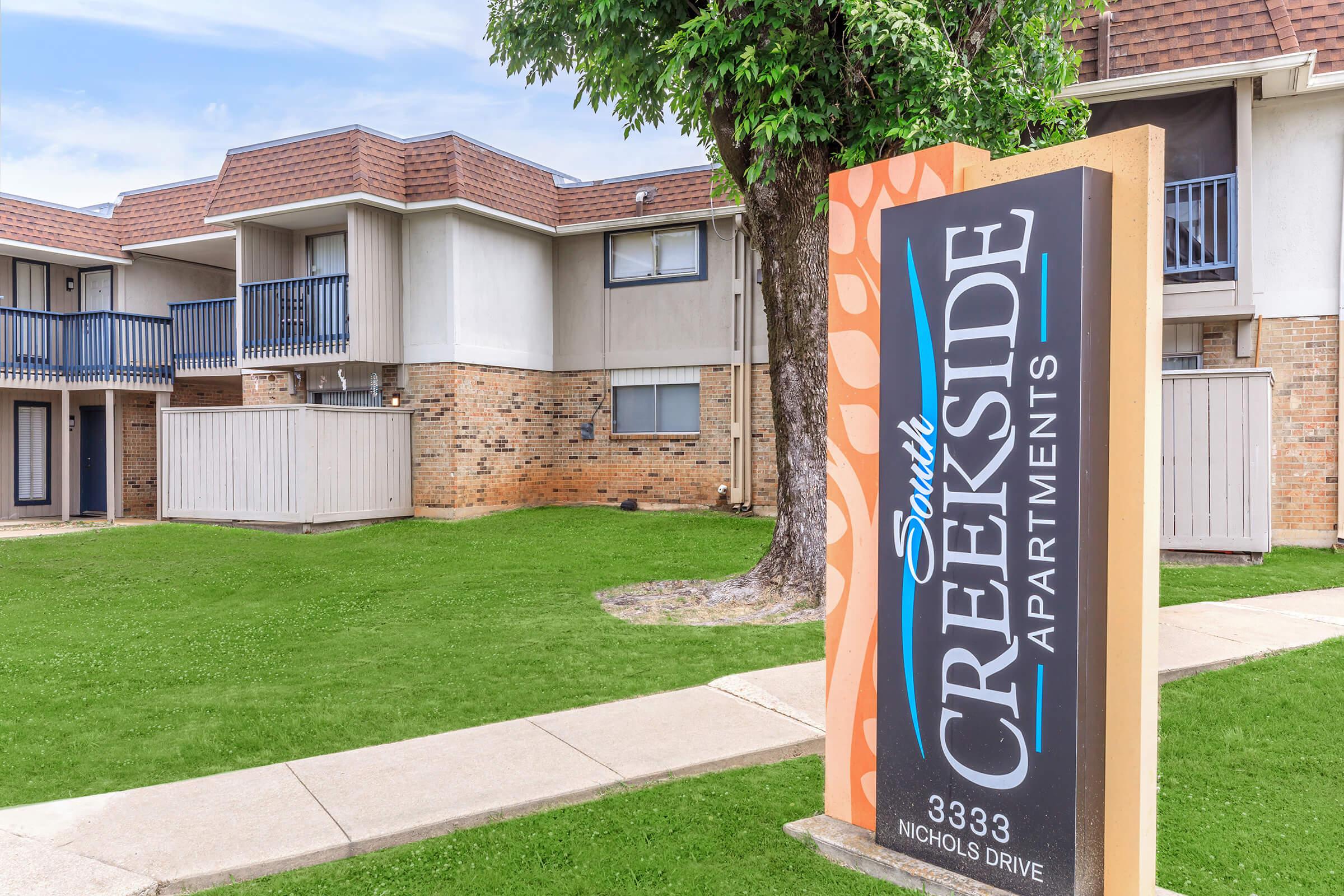 A sign for South Creekside Apartments prominently displayed at the entrance, with the building complex and green lawn in the background. The apartments have balconies and a brick facade, surrounded by trees and walkways.