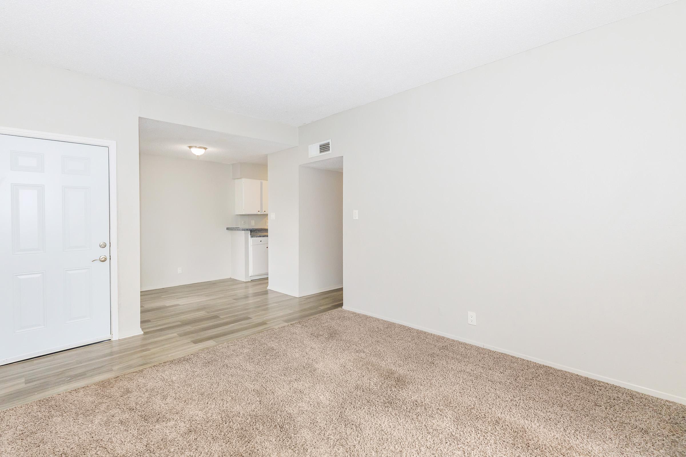 A spacious living area with light beige carpeting and neutral-colored walls. A white front door is visible on the left, and an open doorway leads to a kitchen area in the background, featuring light cabinetry. The overall ambiance is bright and inviting, with natural lighting highlighting the space.