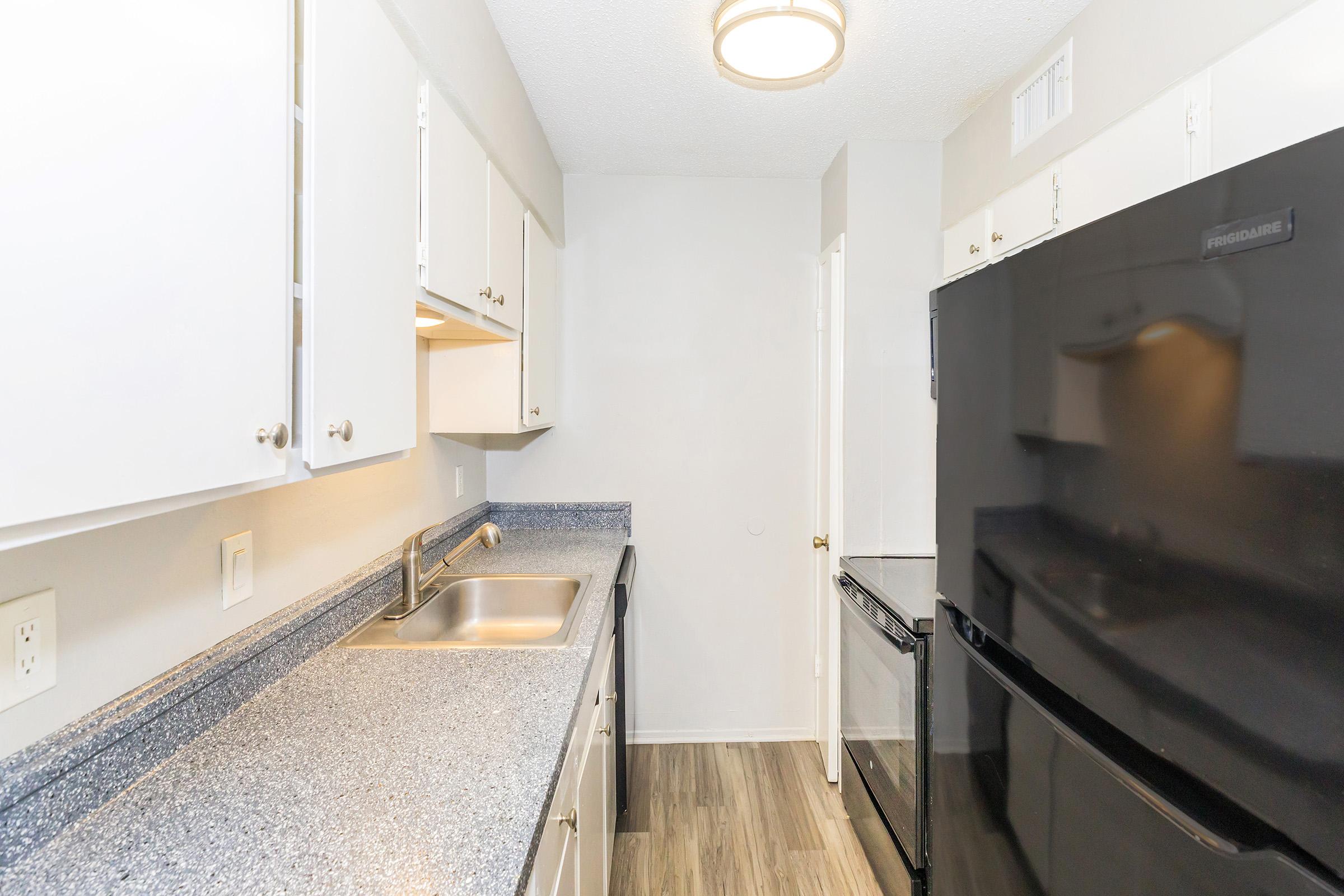 A compact kitchen featuring white cabinetry, a double sink, and modern black appliances including a refrigerator and stove. The countertops are a light gray color, and the room has a neutral wall color with wood-like flooring, creating a clean and functional cooking space.