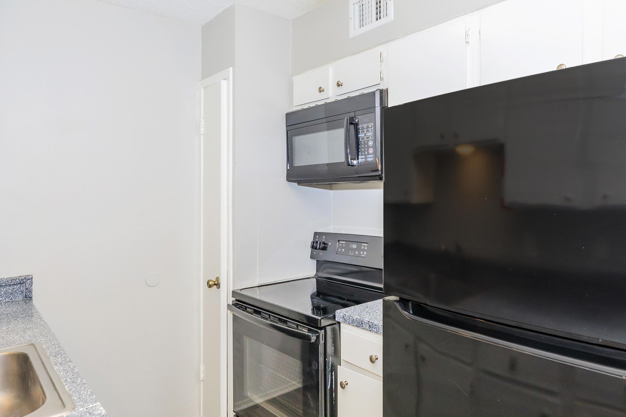 A kitchen area featuring a black refrigerator, microwave, and stove. The cabinets are white, and the countertop has a speckled grey finish. A door leads to another room or area, and a sink is visible in the corner. The walls are light-colored, creating a modern and clean aesthetic.