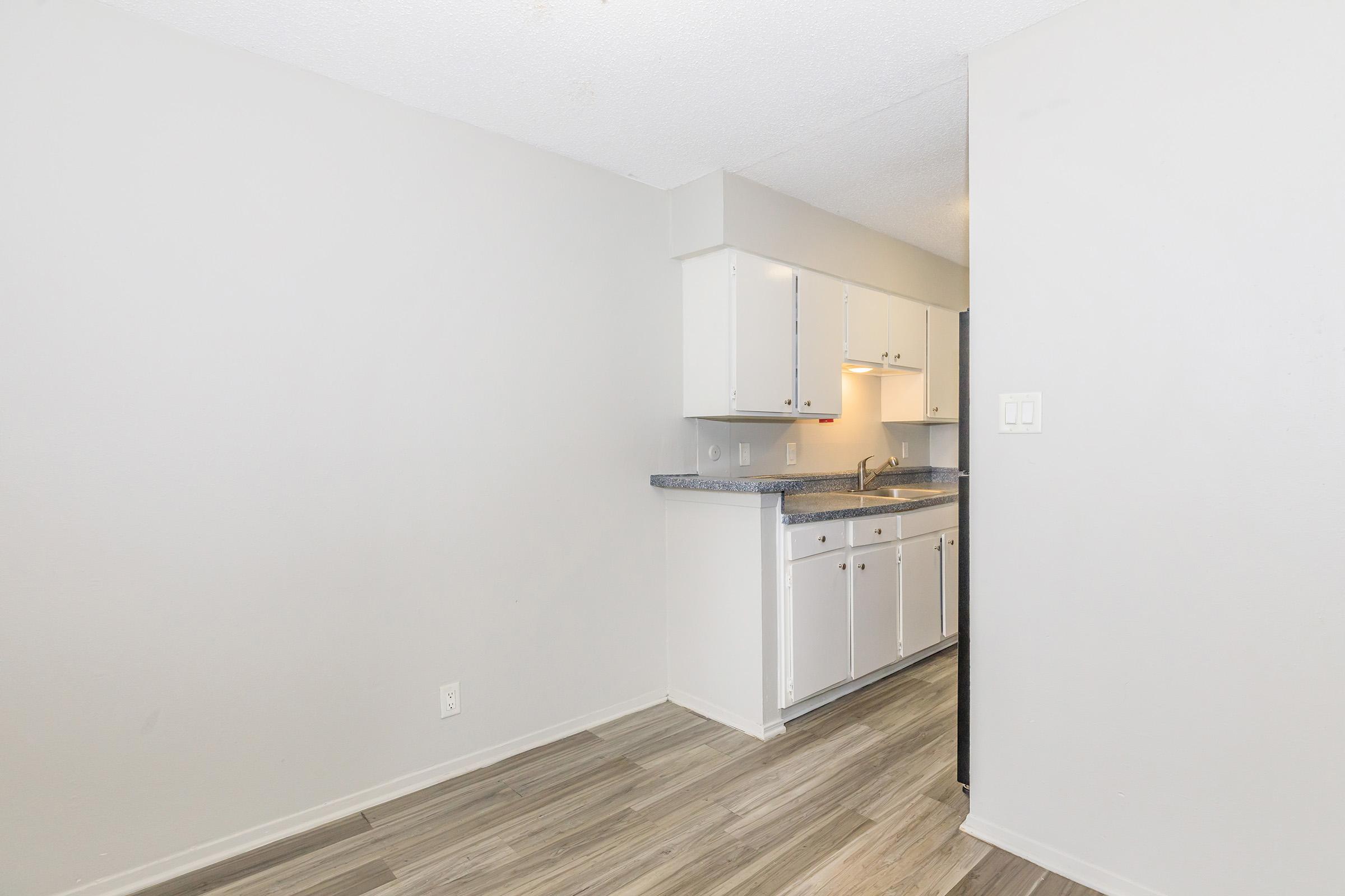 A small, modern kitchen with white cabinets and a gray countertop. The floor features light wood-like tiles. The kitchen area includes a sink and some appliances, with a white wall in the background, creating a clean and minimalistic look.