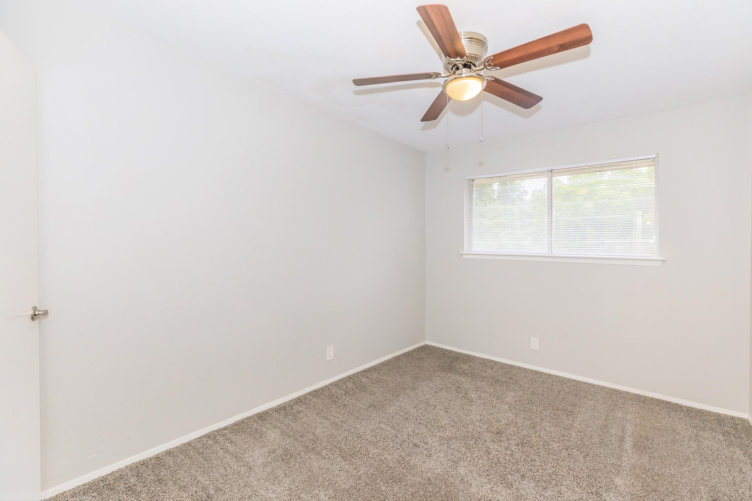 A bright, empty room with light gray walls and carpet. A ceiling fan with wooden blades is installed, and a window with blinds provides natural light. The space is minimally decorated, ideal for various uses or personal customization.