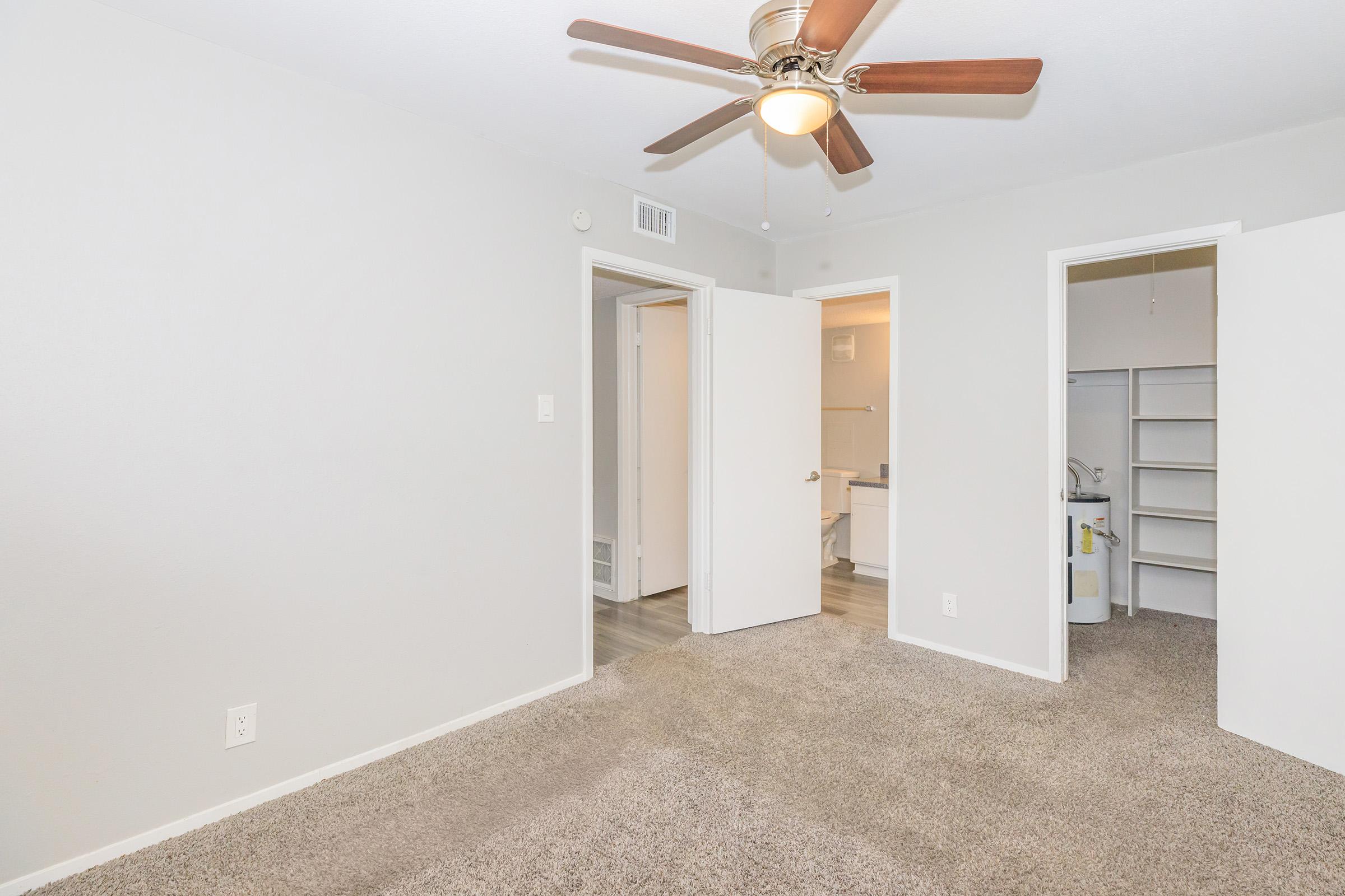 Interior of a light-colored room featuring a ceiling fan with wooden blades. The floor is carpeted in a neutral tone. Two doorways lead to adjoining rooms, one of which has a closet with shelves. The overall appearance is clean and modern, suitable for a living space.