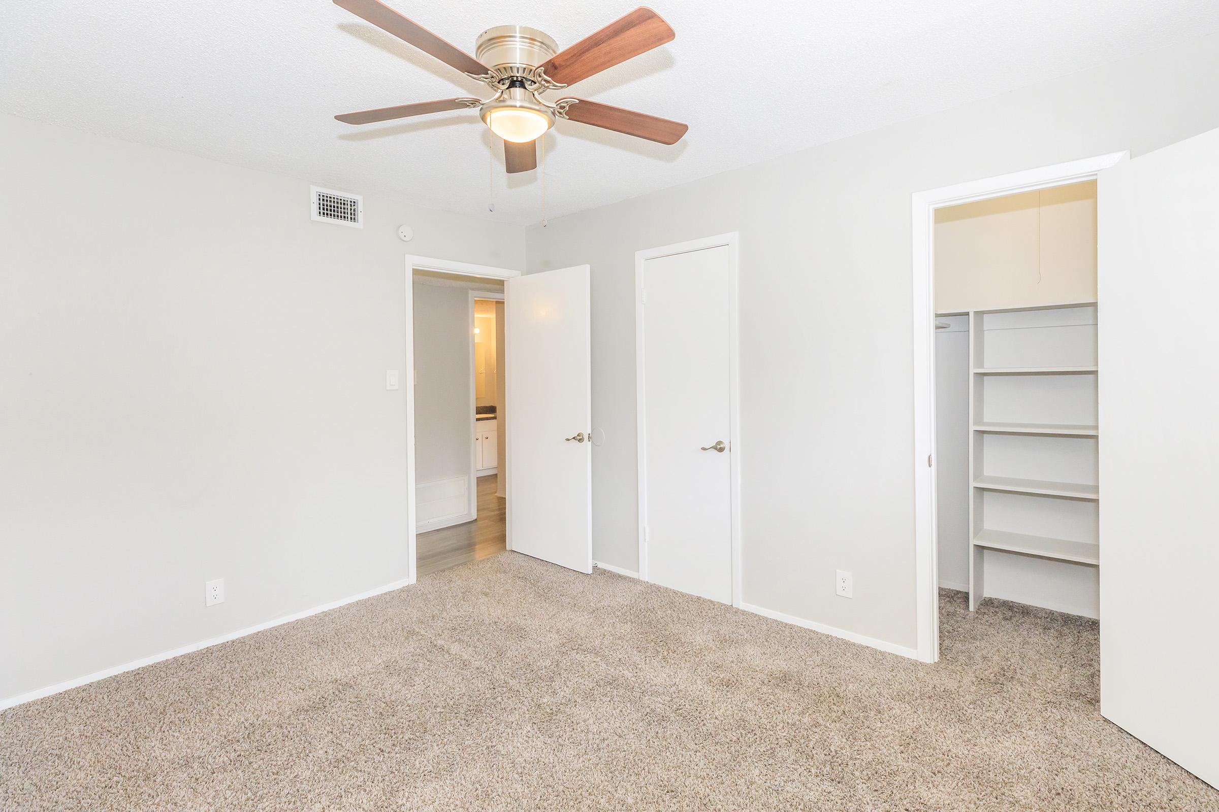 A spacious room with light-colored walls and carpet. It features a ceiling fan, two white doors leading to closets, and natural light coming from a nearby window. One door opens to a bathroom area, creating a bright and airy feel in the space.