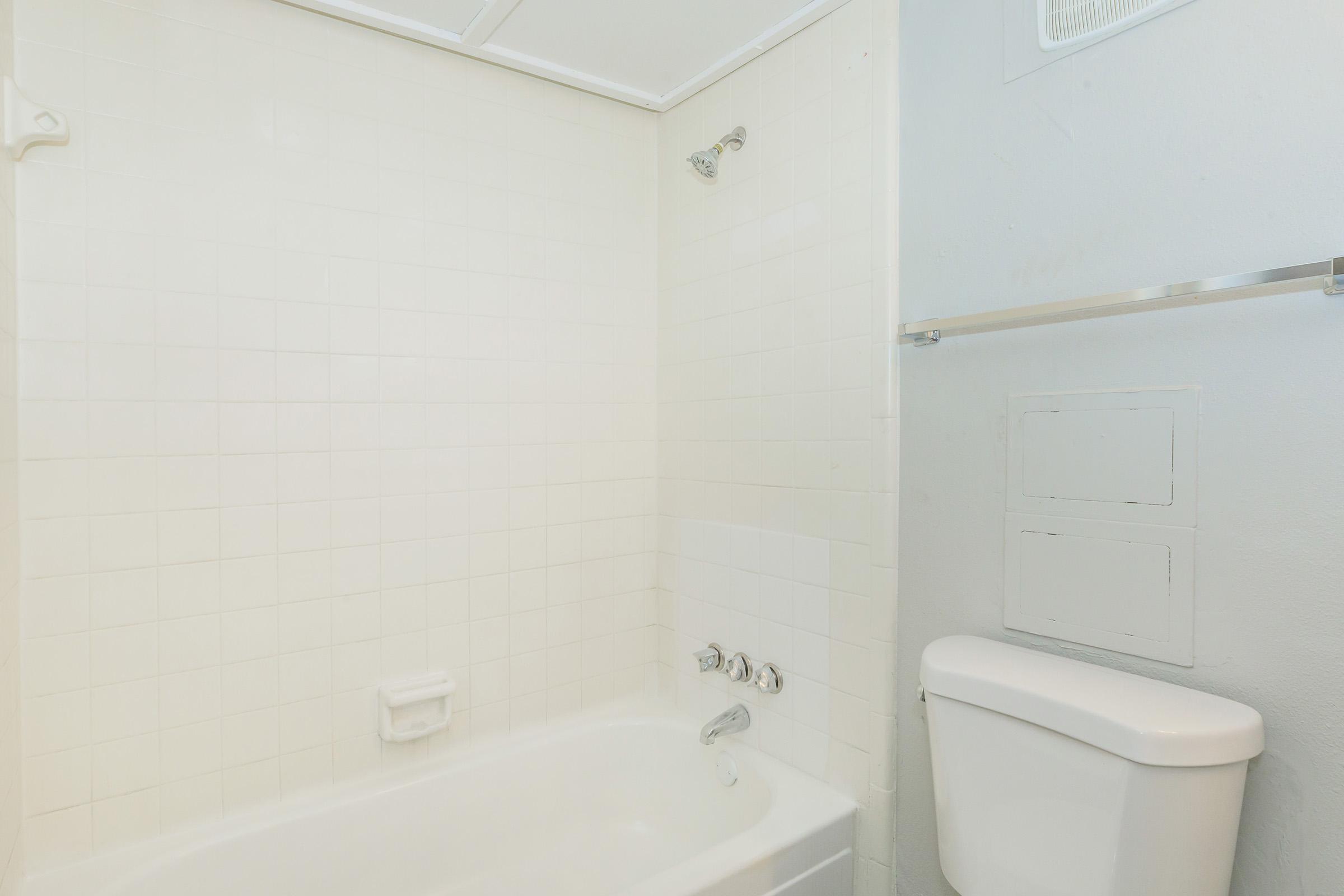 A clean, simple bathroom featuring a white bathtub with a showerhead, white tiled walls, a toilet, and a towel bar. The space is well-lit, with a neutral color scheme and minimal decor, emphasizing a fresh and tidy appearance.