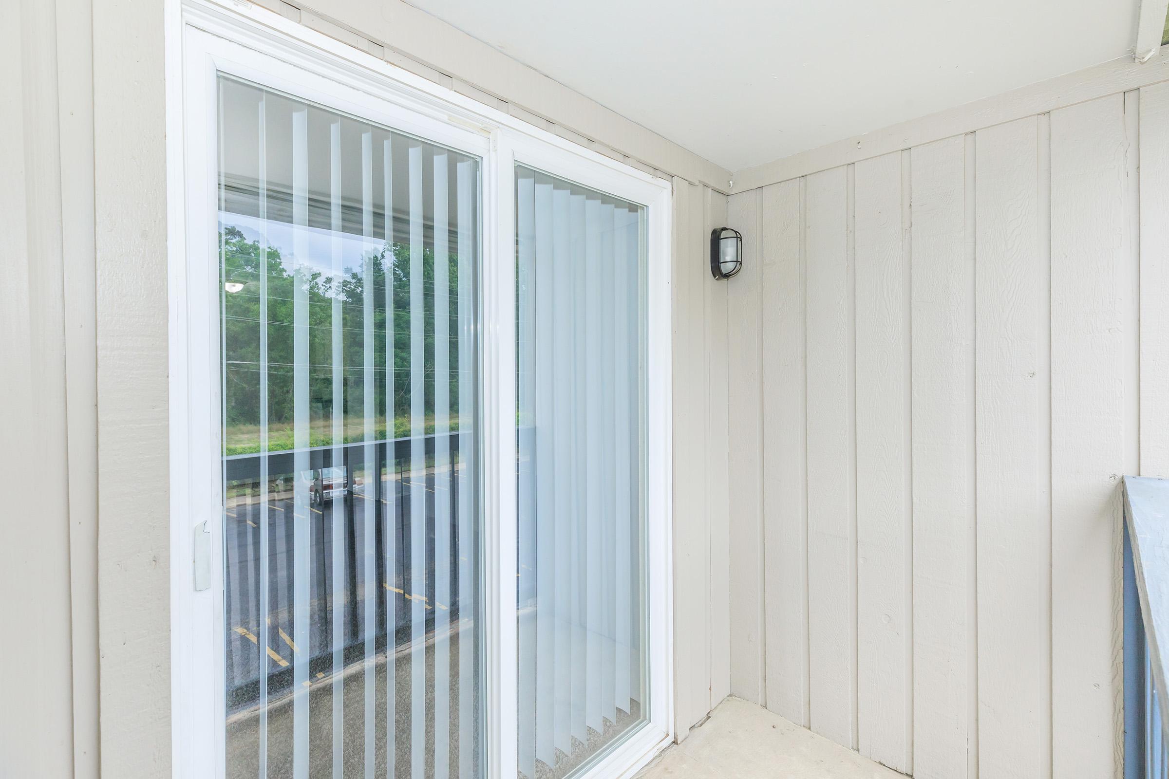 A view of a small balcony area with beige horizontal wooden siding. The balcony features sliding glass doors with vertical blinds, allowing natural light to enter. A light fixture is mounted on the wall beside the doors, and the background shows a parking lot and greenery beyond.
