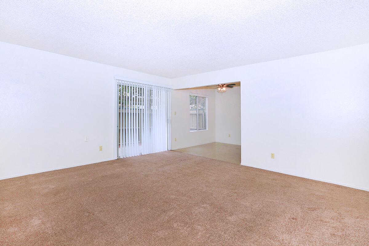 Empty living room with beige carpet and white walls. A sliding glass door is visible, allowing natural light, and leading to an outdoor area. The room features a ceiling fan and a doorway that connects to another room. The space appears clean and ready for furnishing.