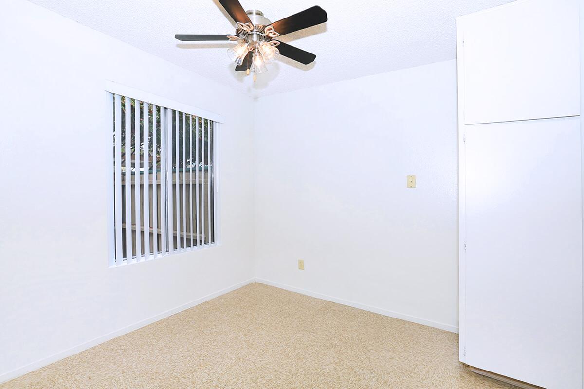 A brightly lit, empty room featuring a ceiling fan with five blades, white walls, and a window with vertical blinds. The floor is covered with beige carpeting. A small white closet is visible on the right side. Overall, the space appears clean and ready for furniture.