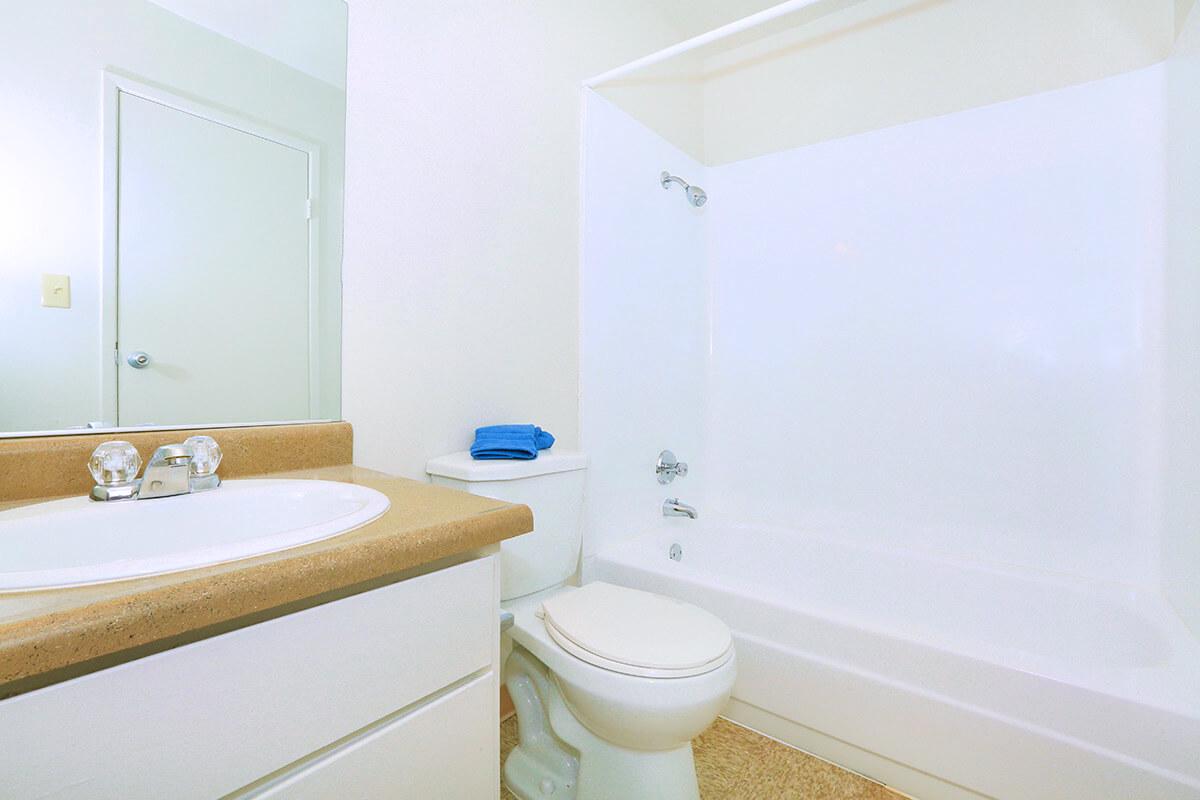 A clean, modern bathroom featuring a white bathtub and shower, a toilet beside it, and a sink with a beige countertop. A blue towel is neatly arranged on the edge of the tub. The walls are painted a light color, creating a bright and airy atmosphere. A closed door is visible in the background.