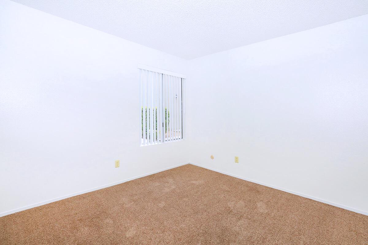 Empty room with beige carpet and white walls. A small window with vertical blinds allows natural light, creating a bright atmosphere. The space is void of furniture, emphasizing its potential for personalization and decoration.