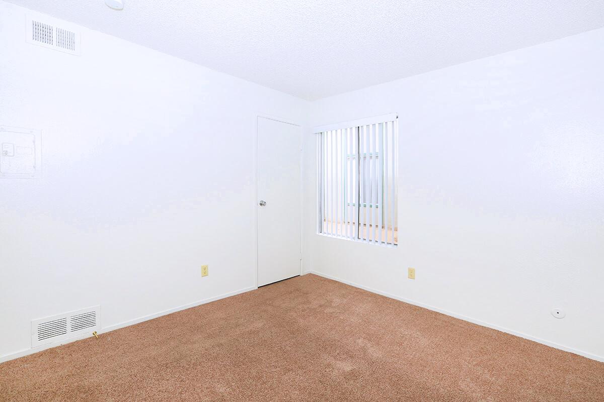 A vacant room with light-colored walls and tan carpet. There is a window with vertical blinds and a door leading to another area. The space is empty, creating a bright and open atmosphere.