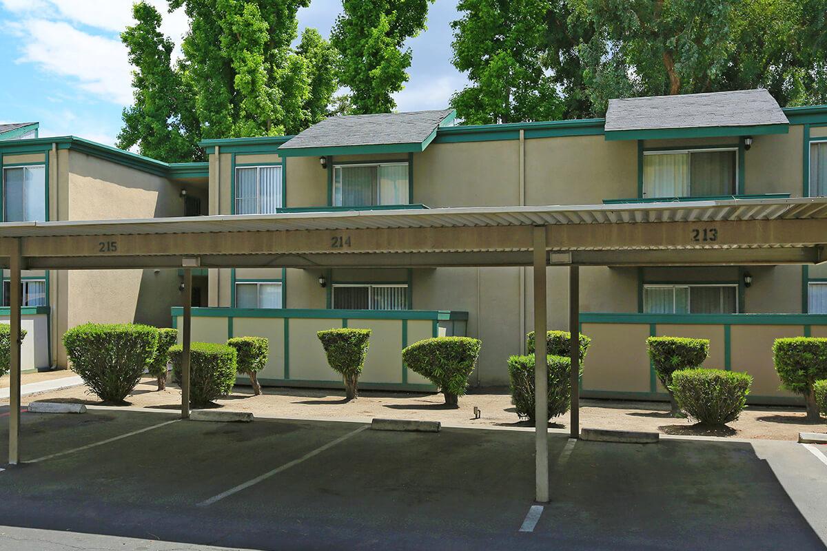 View of a low-rise apartment complex featuring several units with green trim. The foreground shows covered parking spaces with numbers 213, 214, and 215 visible. Neatly trimmed bushes line the parking area, and there are trees in the background under a clear blue sky.