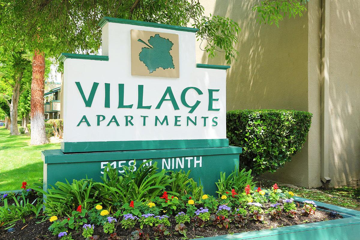 Sign for Village Apartments featuring a green and white color scheme, with the words "VILLAGE APARTMENTS" prominently displayed. Below the main sign is the address "515 E NINTH" and a small decorative map outline. The base of the sign includes colorful flowerbeds and greenery.