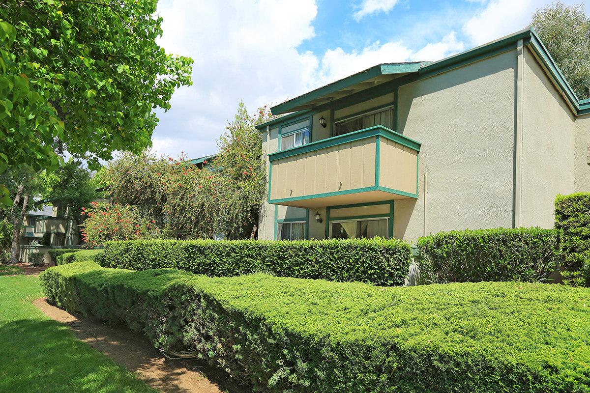 Two-story apartment building surrounded by well-maintained shrubs and grass. The building features a balcony on the upper level and is set against a backdrop of blue sky with scattered clouds. Lush greenery enhances the inviting outdoor space.