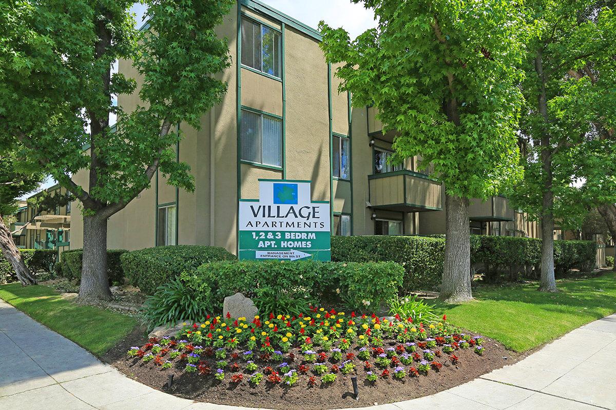 A view of Village Apartments featuring a large sign displaying "Village Apartments 1, 2, 3 Bedroom Apt. Homes." The building has multiple stories with balconies, surrounded by neatly trimmed hedges and a colorful flower bed in front, creating a welcoming atmosphere.
