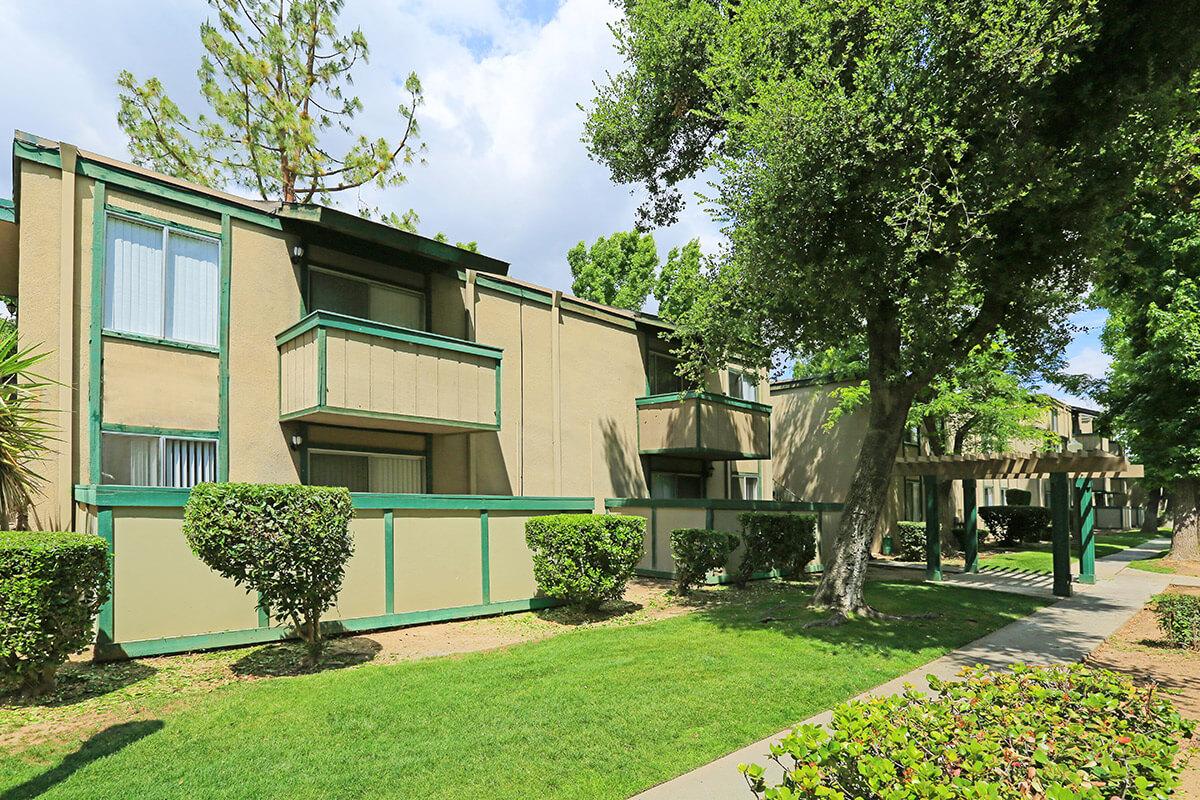 Two-story apartment building with green trim, surrounded by well-maintained landscaping, including shrubs and trees. The building features balconies and multiple windows, with a pathway leading to a shaded area with a pergola. The scene is bright, with blue skies and a few clouds.