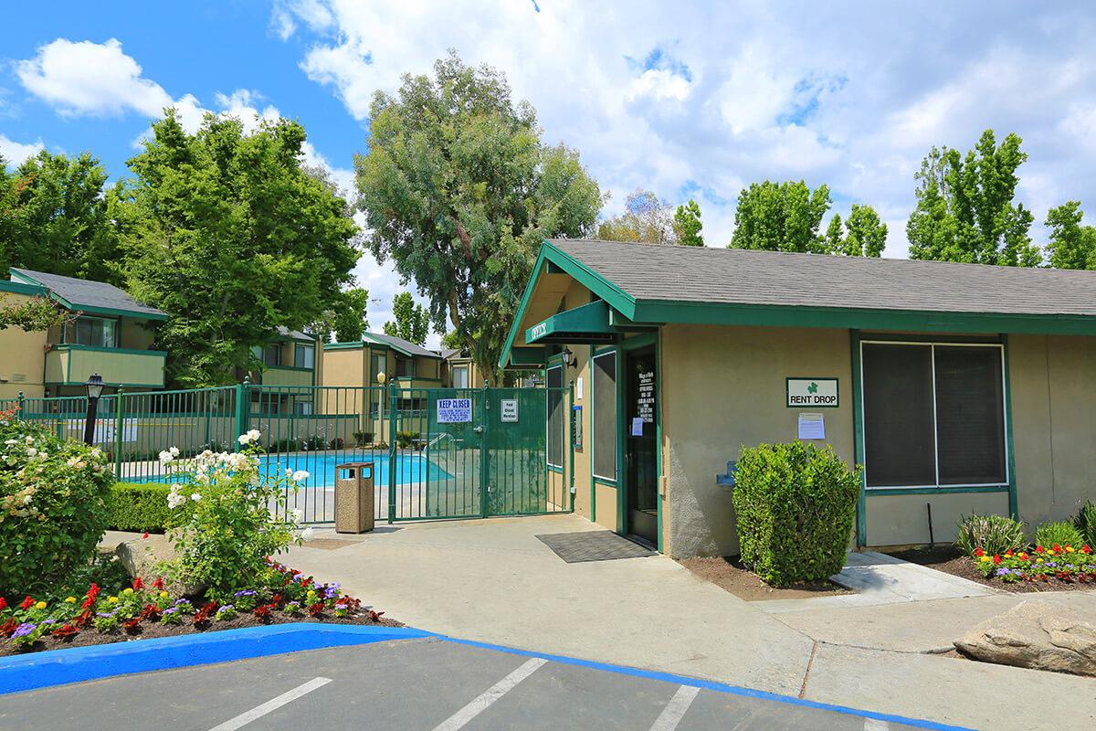 A view of an apartment complex entrance featuring a rental office, a swimming pool visible behind a green fence, and landscaped flower beds. The sky is partly cloudy, and there are trees in the background, adding to the serene atmosphere of the community.