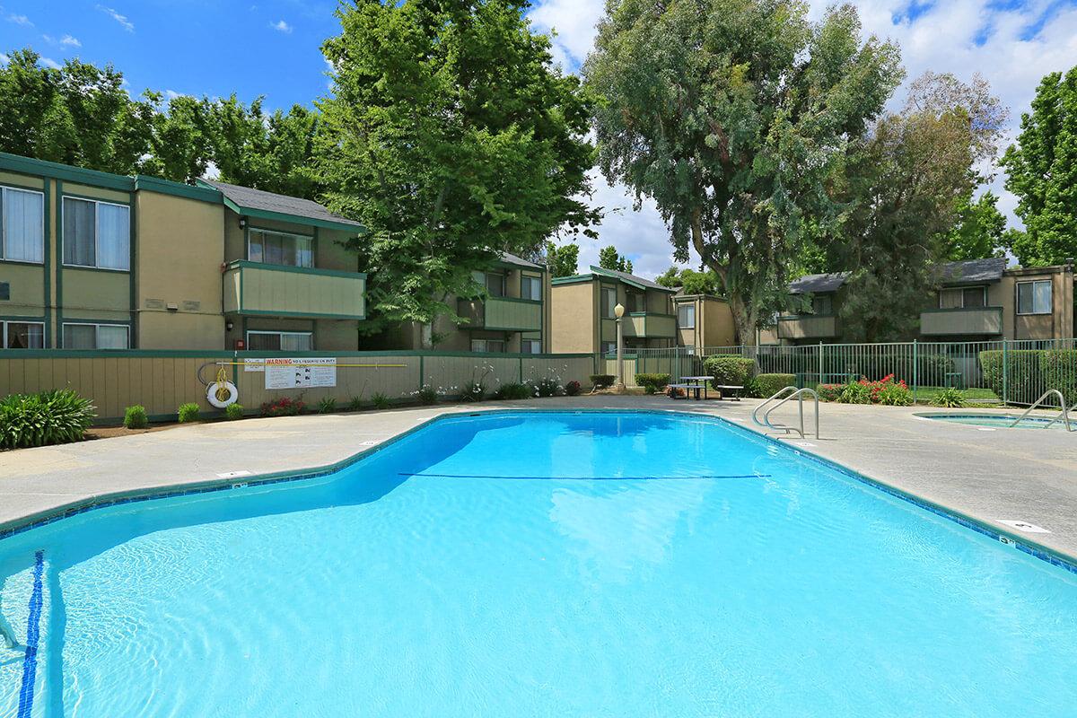 A clear blue swimming pool surrounded by lush greenery and residential buildings. The pool area features lounge chairs and is enclosed by a fence. Trees and shrubs add to the inviting atmosphere under a bright blue sky.