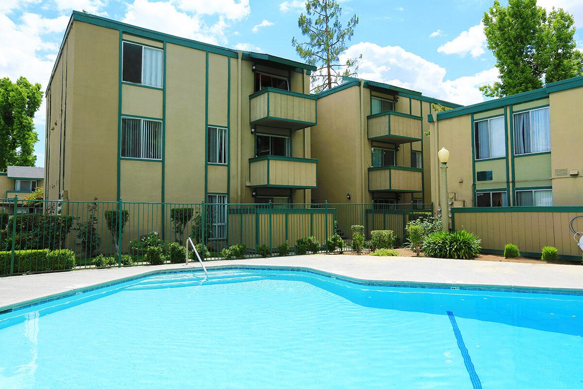 A view of an apartment complex with two-story buildings surrounding a clear blue swimming pool. Lush greenery and trees are visible in the background, under a bright, partly cloudy sky. The pool area features a diving board and is enclosed by a decorative fence.
