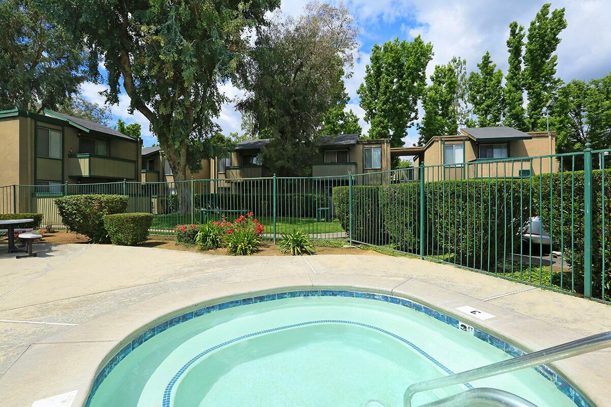 A view of a hot tub in the foreground with a landscaped area and green bushes. In the background, there are several two-story residential buildings surrounded by trees and a fence. The sky is partly cloudy, creating a serene outdoor setting.