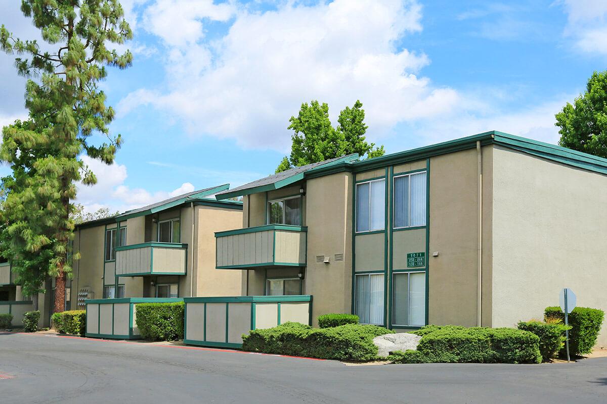 A modern two-story apartment complex with green accents. The building features multiple balconies, large windows, and is surrounded by well-maintained shrubs and trees. The sky is bright with fluffy clouds, creating a pleasant atmosphere.