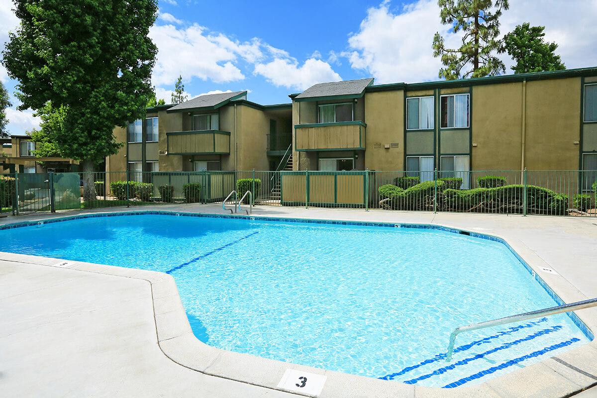 A clear blue swimming pool surrounded by a fenced area, with two-story apartment buildings in the background. Lush green landscaping includes bushes and trees. The sky is partly cloudy, creating a bright and inviting atmosphere.