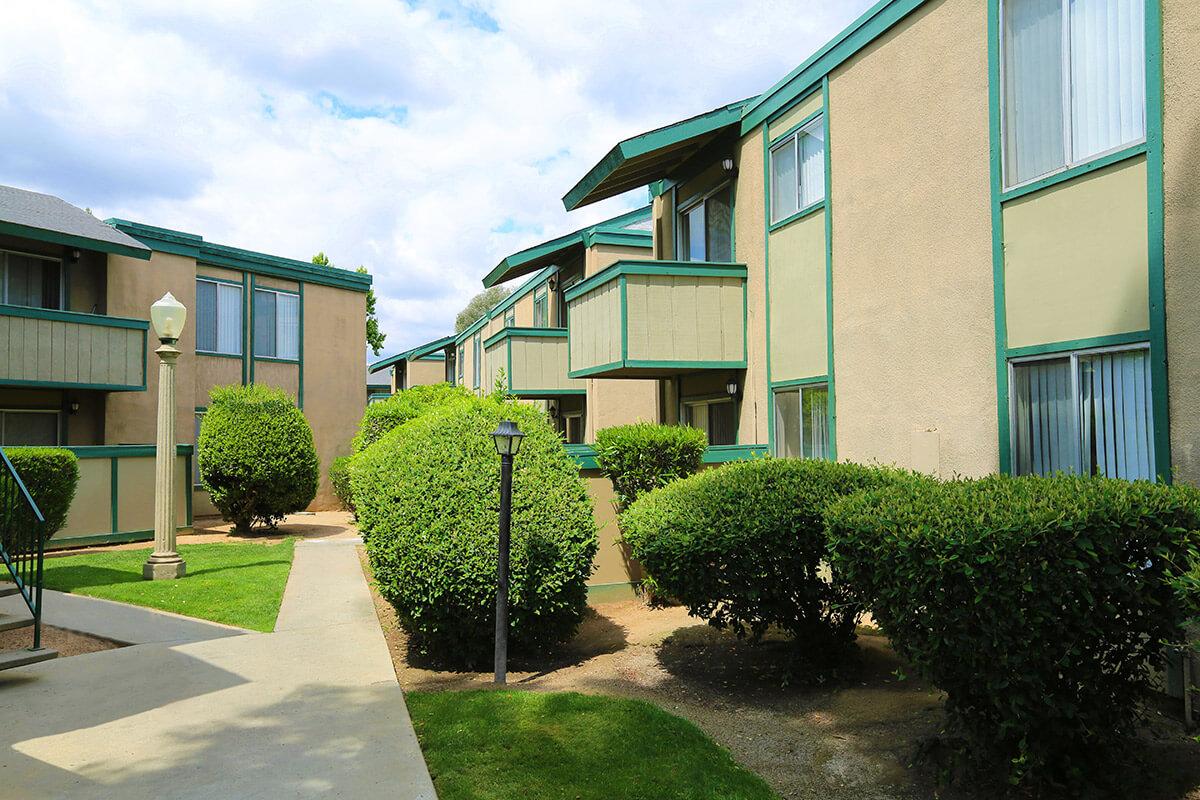 Two-story apartment buildings with green trim and balconies, surrounded by well-maintained shrubs and grass. Pathways lead through the landscaped area, providing access to the units. The sky is partly cloudy, creating a bright, inviting atmosphere.
