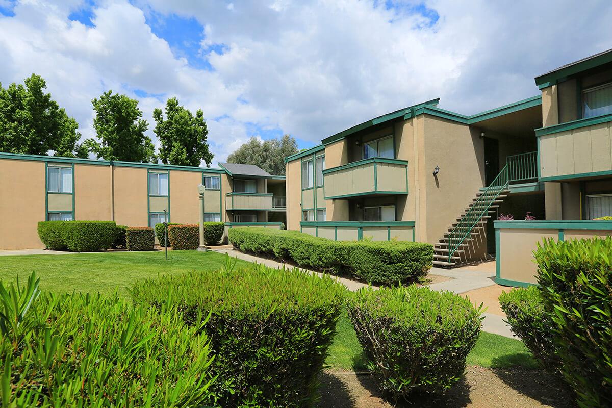 A landscaped area featuring two mid-rise apartment buildings surrounded by greenery. The buildings have balconies and are painted in neutral colors, with a pathway leading to a set of stairs. The sky is partly cloudy, indicating a pleasant day.