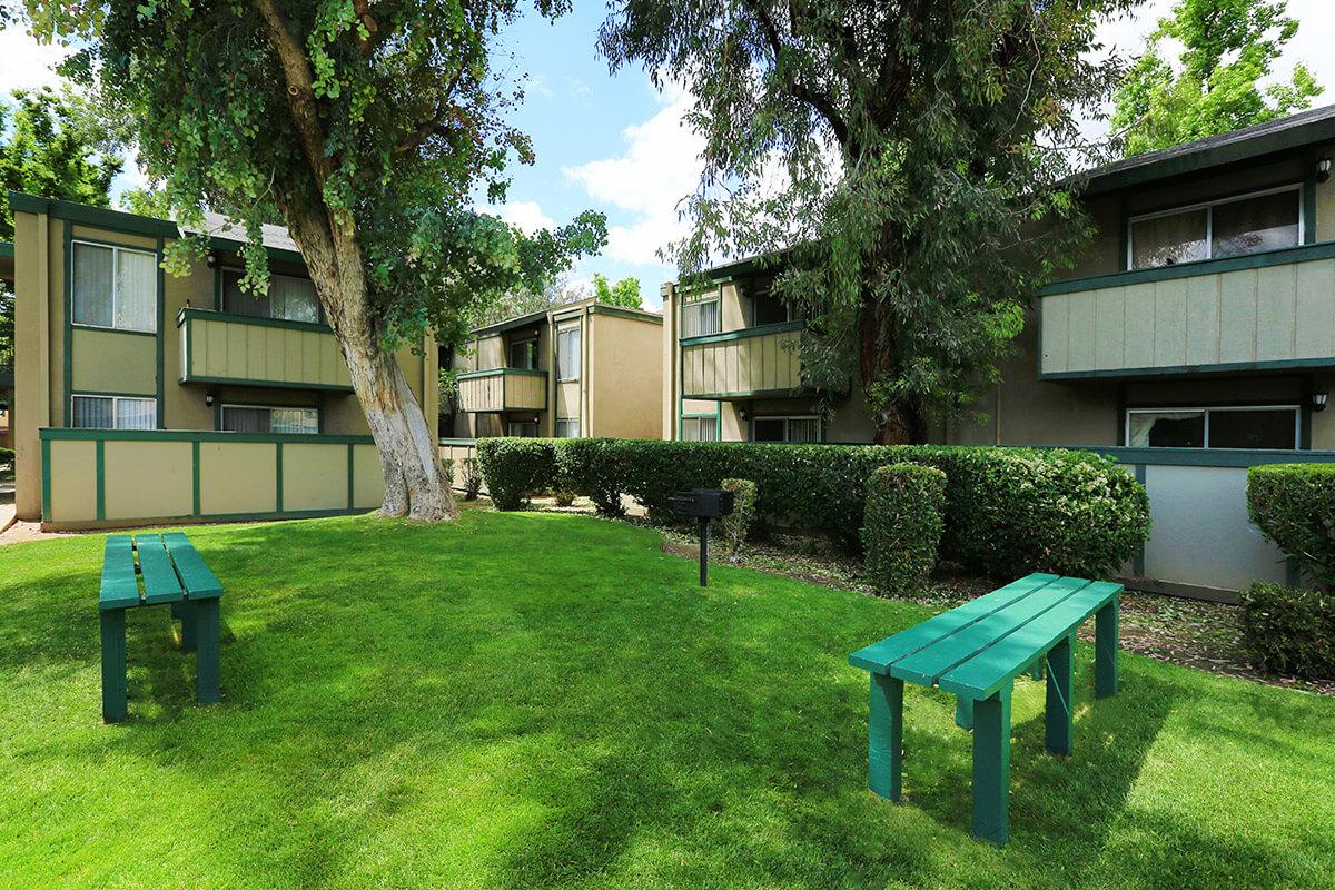View of a grassy area with two green benches in the foreground, surrounded by well-maintained shrubs. In the background, there are three multi-story apartment buildings with balconies and windows under a clear blue sky dotted with clouds. The scene is bright and inviting.