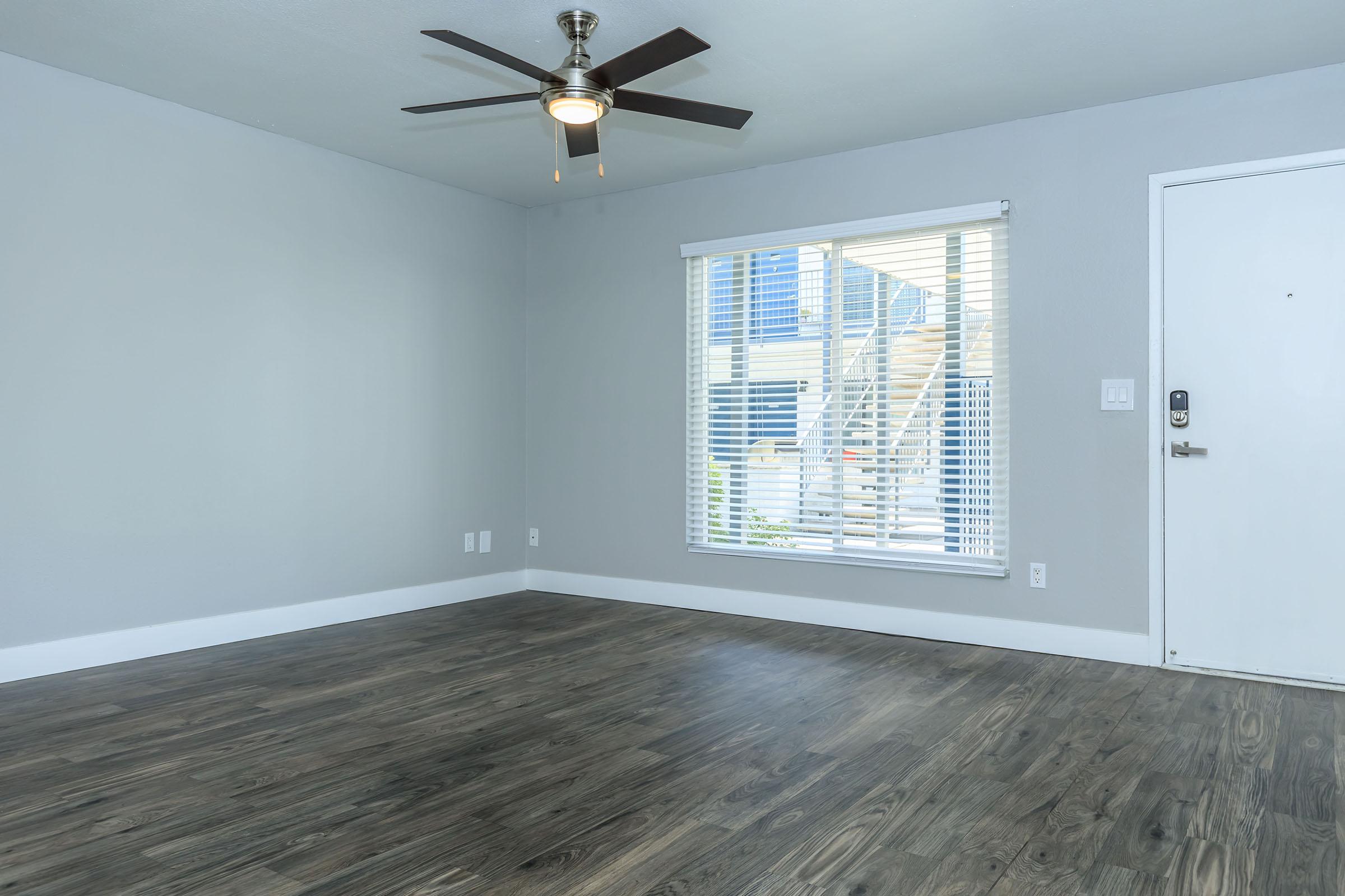 Large living room with brown wooden floors, large front window, and ceiling fan at Rise at the Lofts
