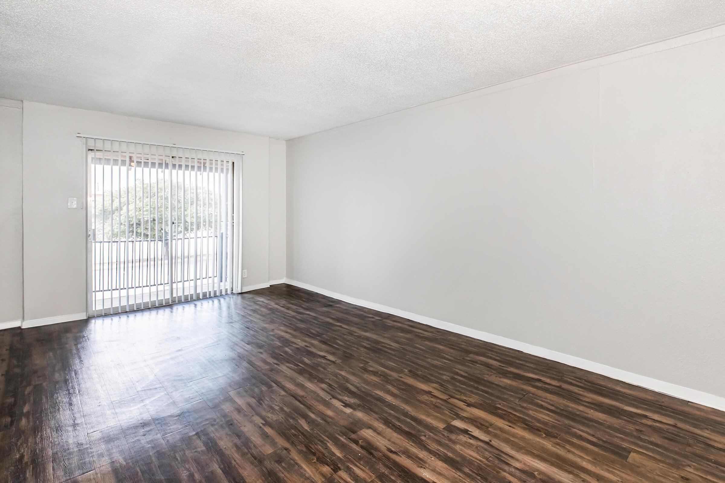 A spacious, empty room with light-colored walls and a wood-like floor. A large window with vertical blinds allows natural light to enter, providing a view of the outdoors. The room appears clean and ready for furnishing.