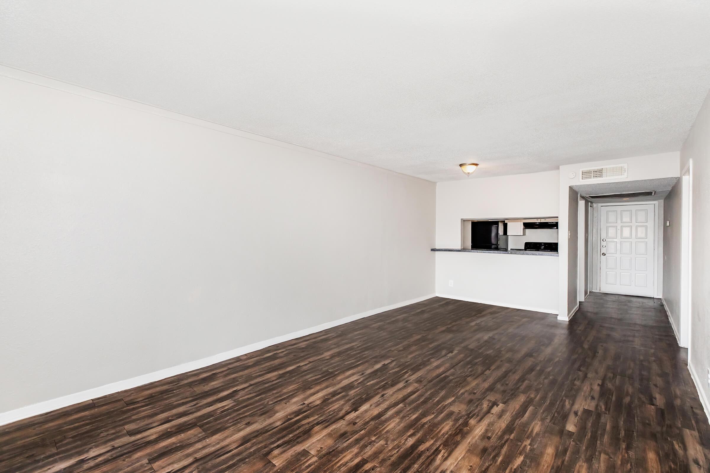 Spacious, empty living area with light-colored walls and dark wood flooring. To the right, a kitchen area is partially visible with a bar counter and black appliances. A doorway leads to another room, creating an open and inviting atmosphere.