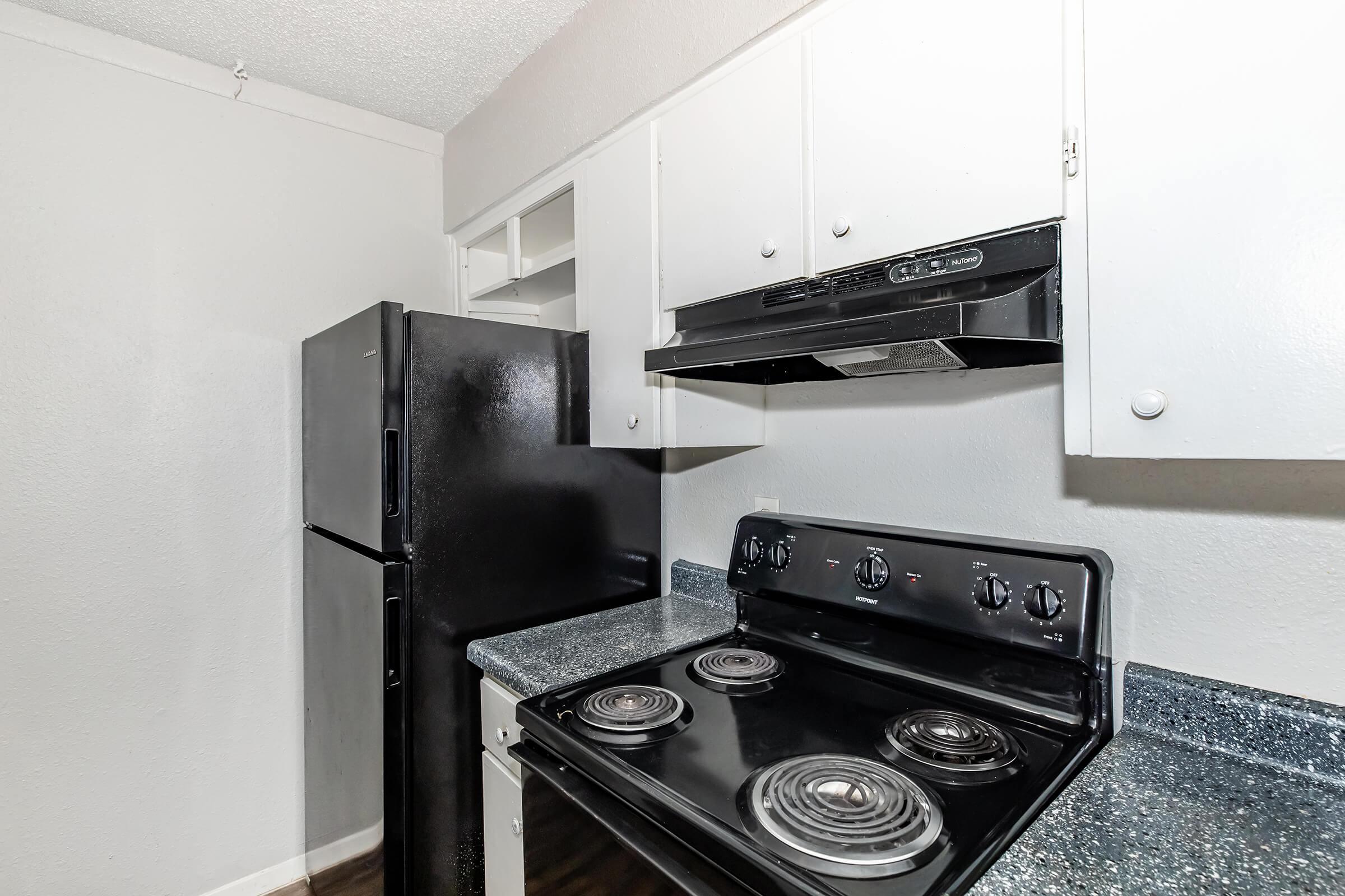 A compact kitchen featuring a black refrigerator, stovetop with four burners, and a range hood. The cabinets above and countertop are light-colored, and the overall color scheme is neutral with gray walls. The kitchen appears clean and functional.