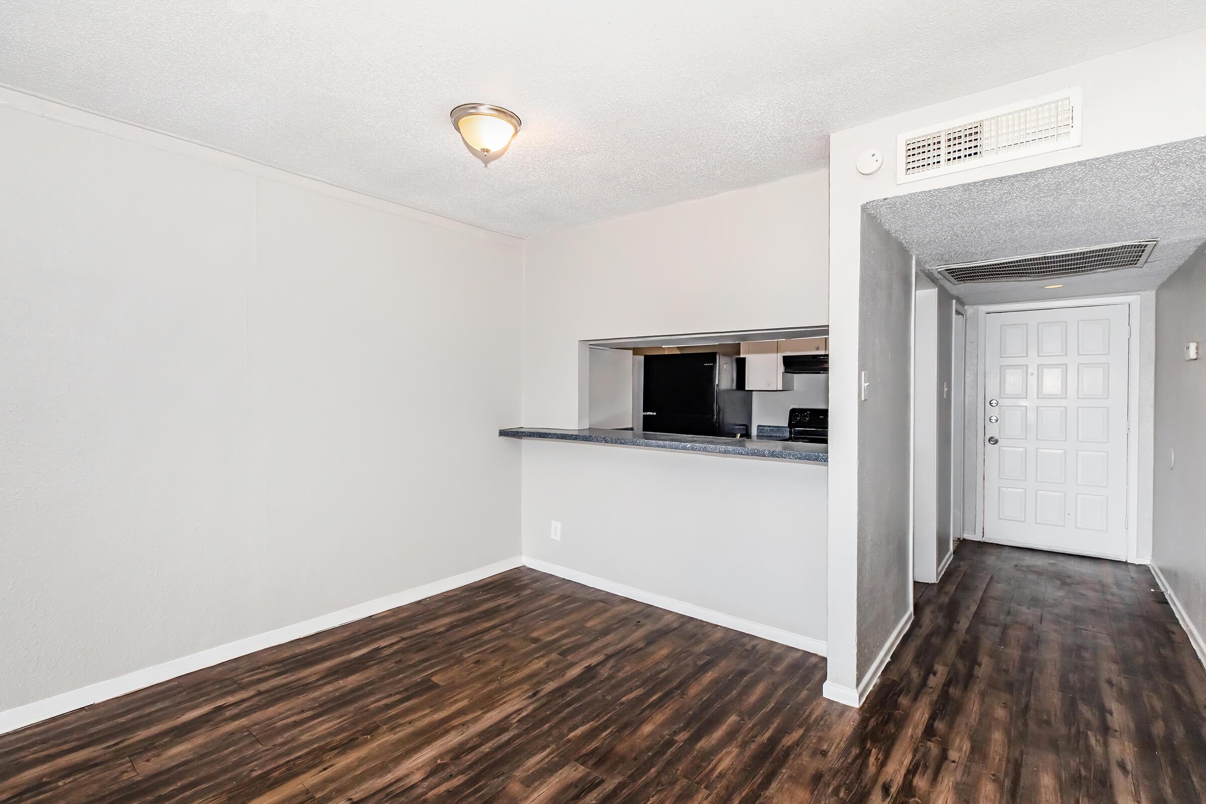 A well-lit living area with a smooth, light gray walls and dark wooden flooring. A small kitchen can be seen in the background through a pass-through opening. The kitchen has modern appliances. To the right, there is a doorway leading to a hallway with a white door, adding to the spacious feel of the room.