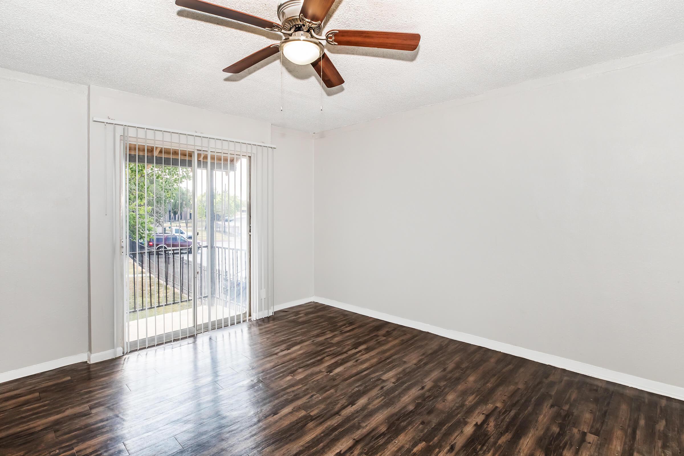A spacious living room with light gray walls and a wooden floor. A ceiling fan with wooden blades is installed. There is a sliding glass door with vertical blinds leading to an outdoor area, allowing natural light to enter. The room is empty, offering a blank canvas for potential decoration.