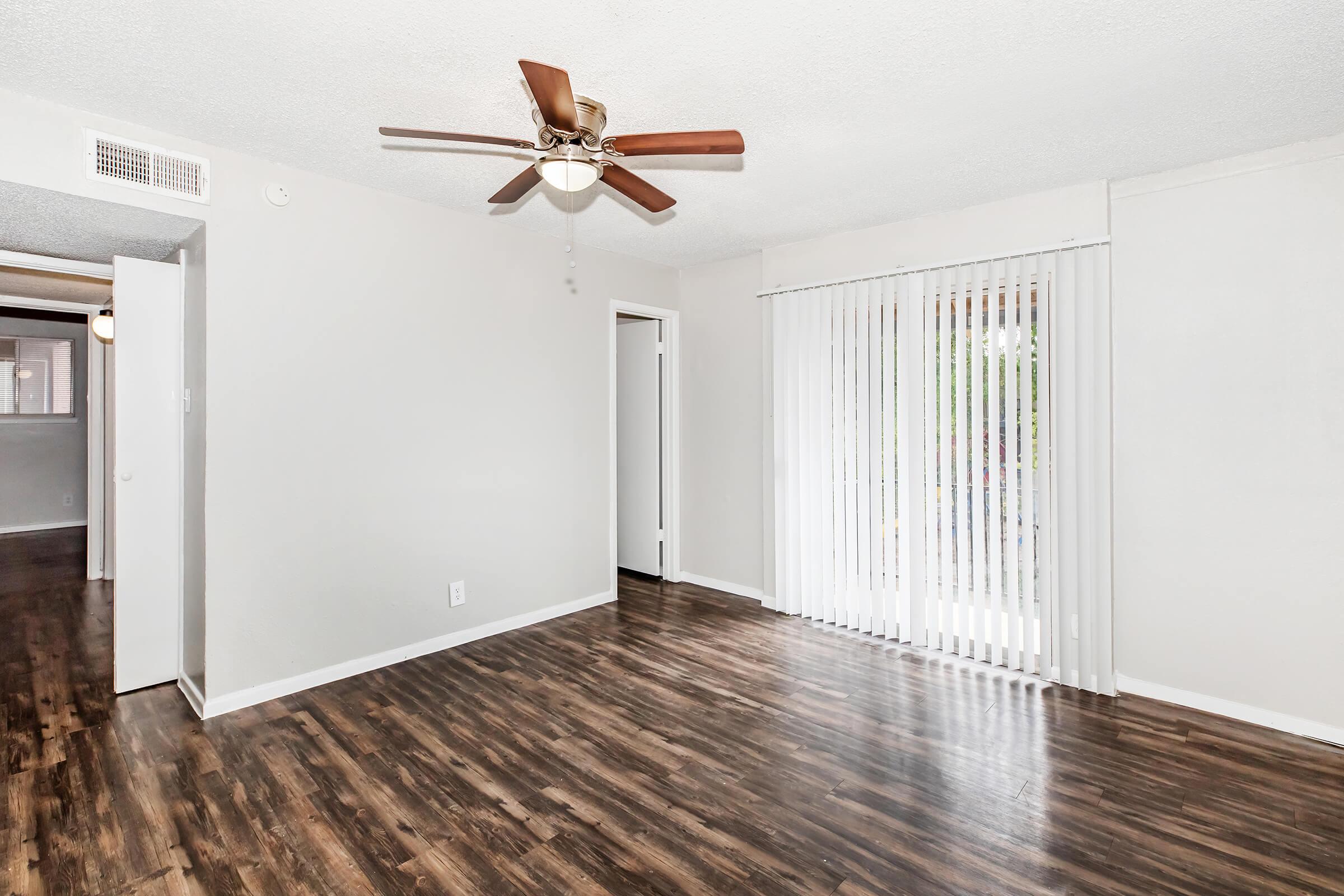 A bright living room with wood-style flooring, a ceiling fan, and large sliding doors covered by vertical blinds. The walls are painted in a light color, enhancing the space's openness. A doorway leads to another room, and the overall atmosphere is inviting and airy.
