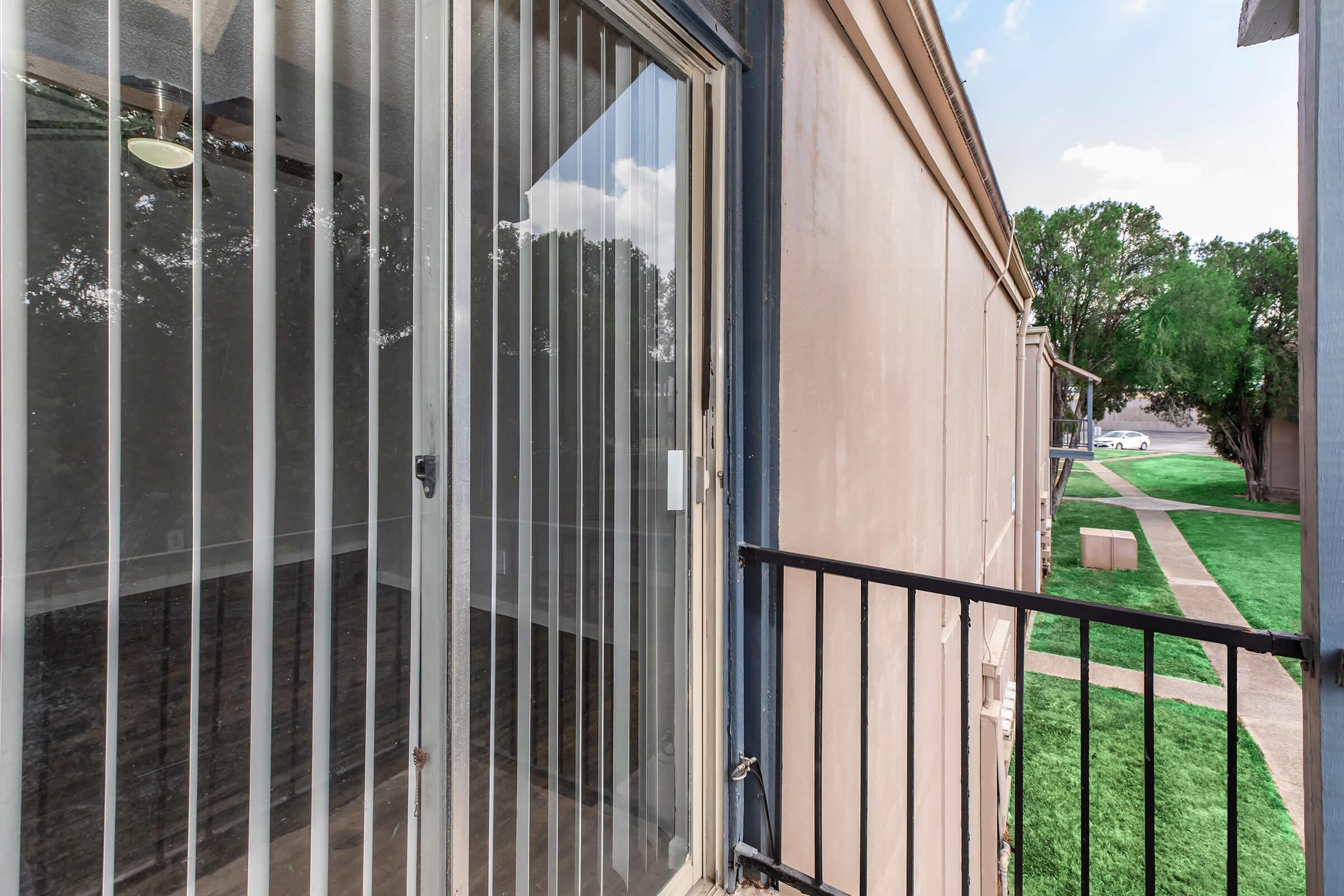 View from a balcony showing vertical blinds, a clear glass door, and a glimpse of a grassy area and pathway outside. The building has a simple exterior with neutral colors and tree foliage in the background.