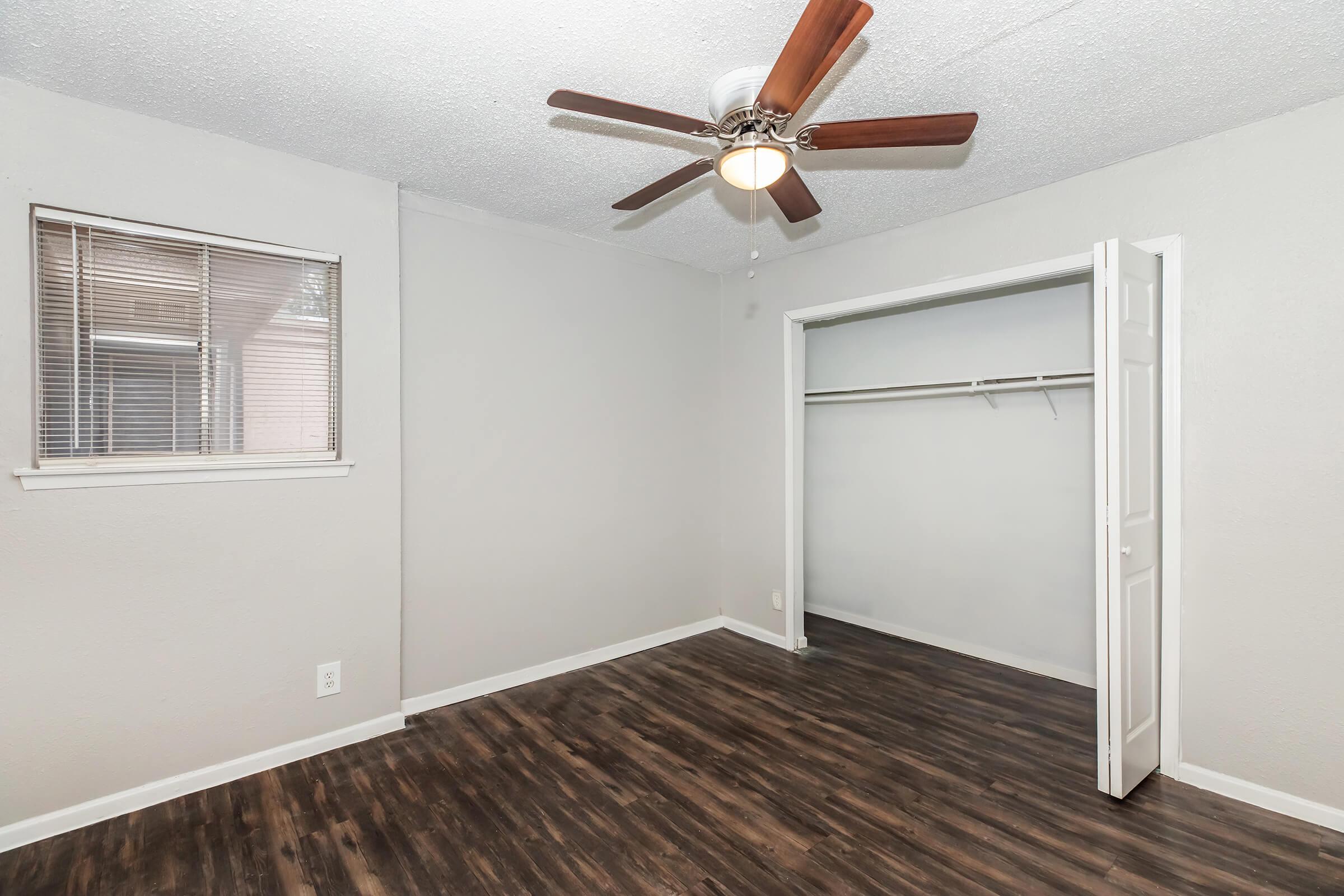 Empty bedroom featuring gray walls, a ceiling fan with wooden blades, and a window. The room has wood-like flooring and a closet with sliding doors. Natural light enters through the window, enhancing the spacious feel of the room.
