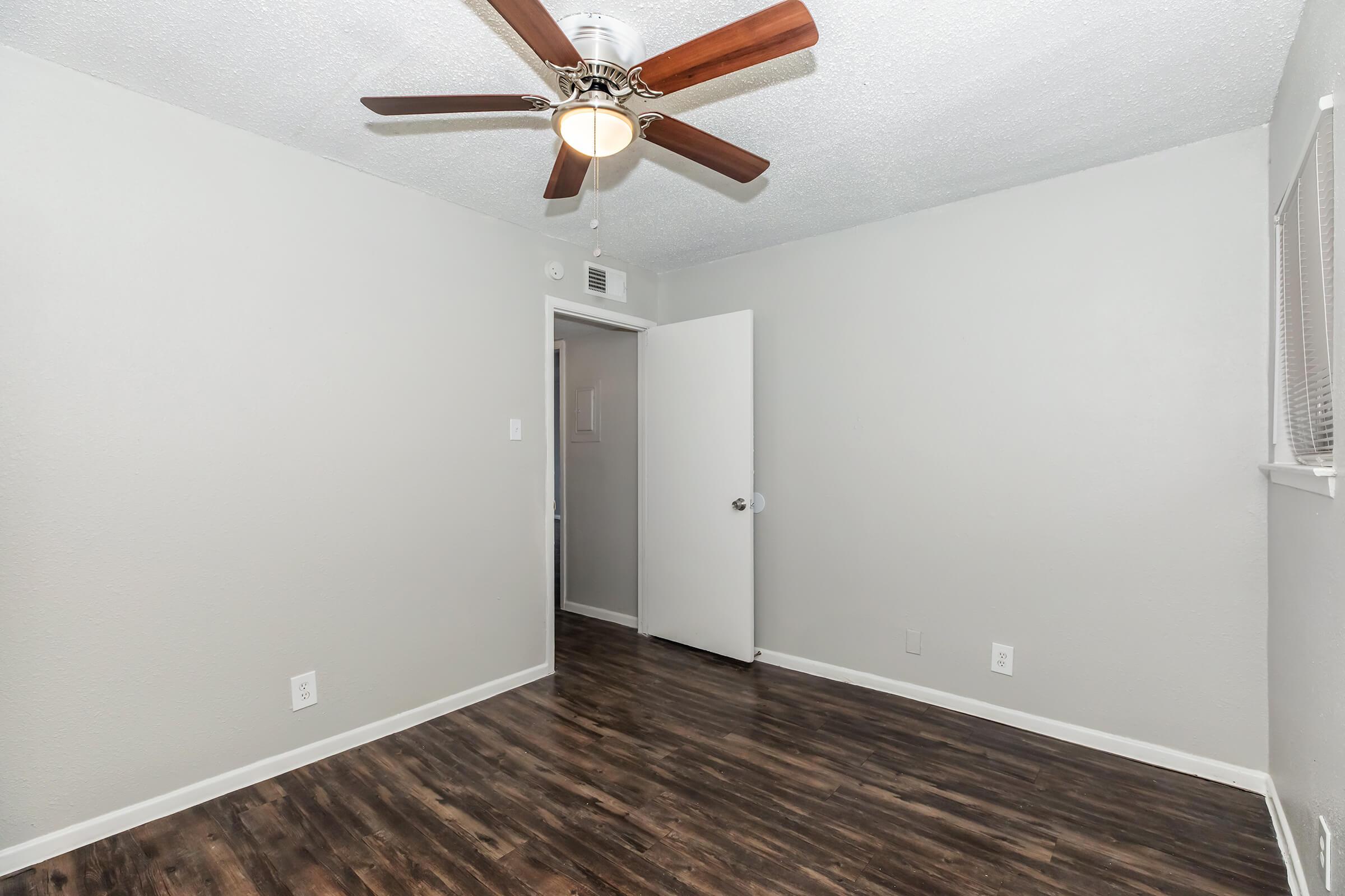 Empty room with light gray walls and a ceiling fan. The floor is dark wood laminate. A closed door is visible on the left, leading to another room or area. A window with blinds is on the right side of the image, providing some natural light.