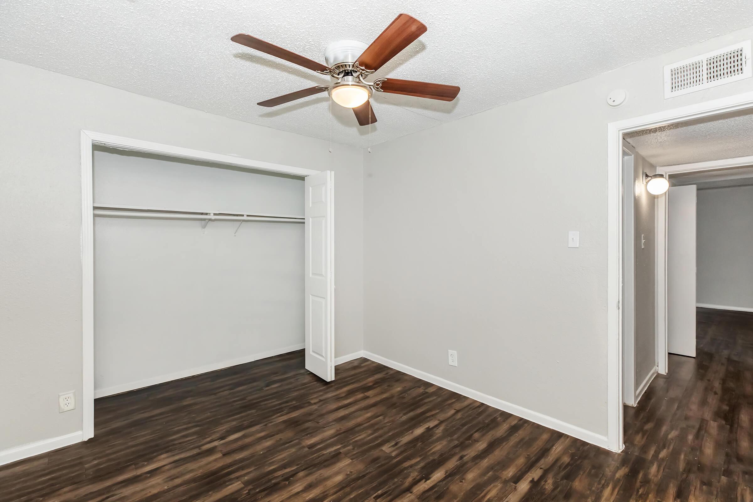 A well-lit room featuring a ceiling fan, light-colored walls, and dark wood flooring. To the left, there is an open closet with a single rod for hanging clothes. A doorway leads to another room on the right, creating a spacious and inviting atmosphere.