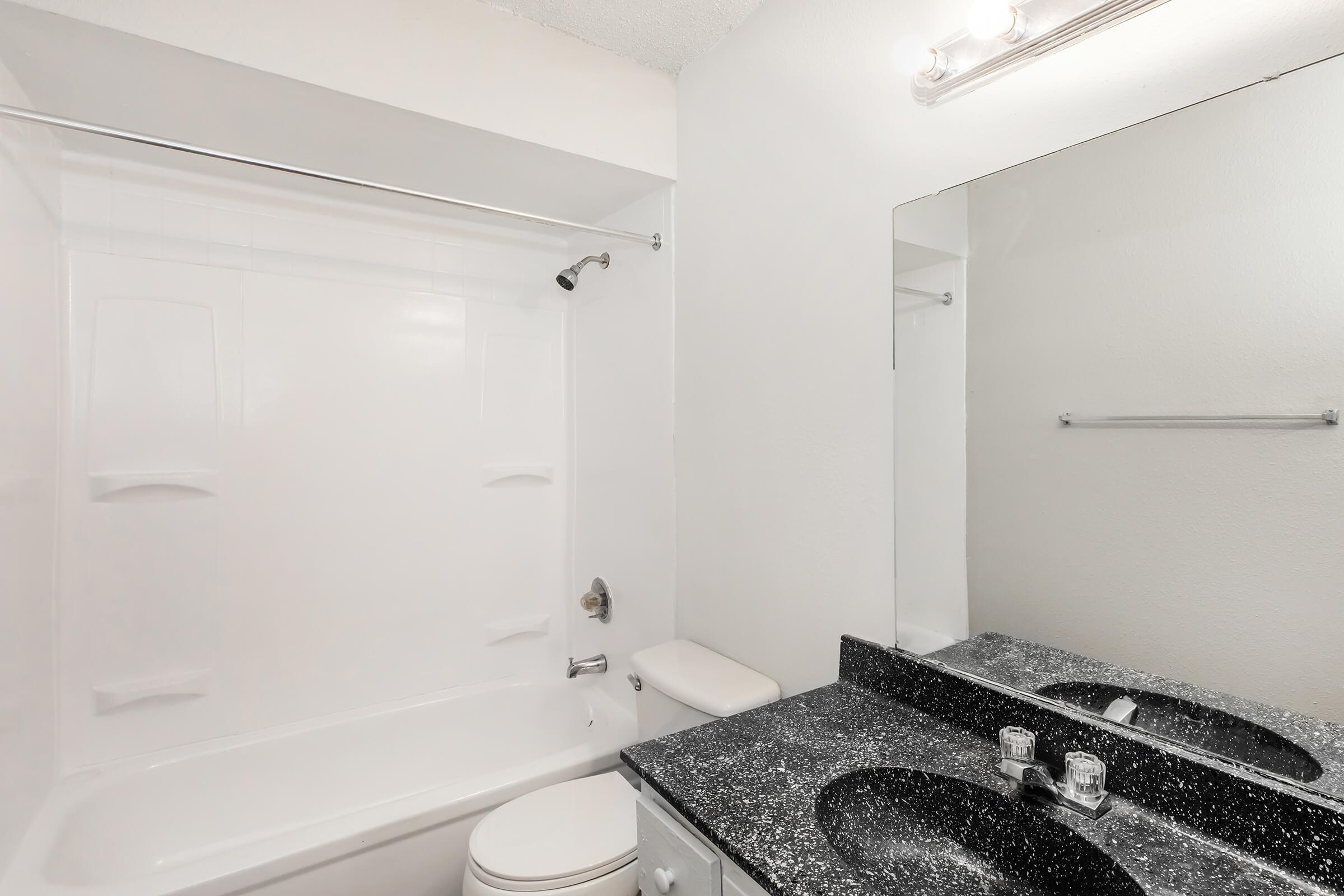 A clean and modern bathroom featuring a white bathtub with a showerhead, a dark speckled countertop with two sinks, and a large mirror above. The walls are painted white, and there is a towel rack next to the mirror. Bright lighting illuminates the space.